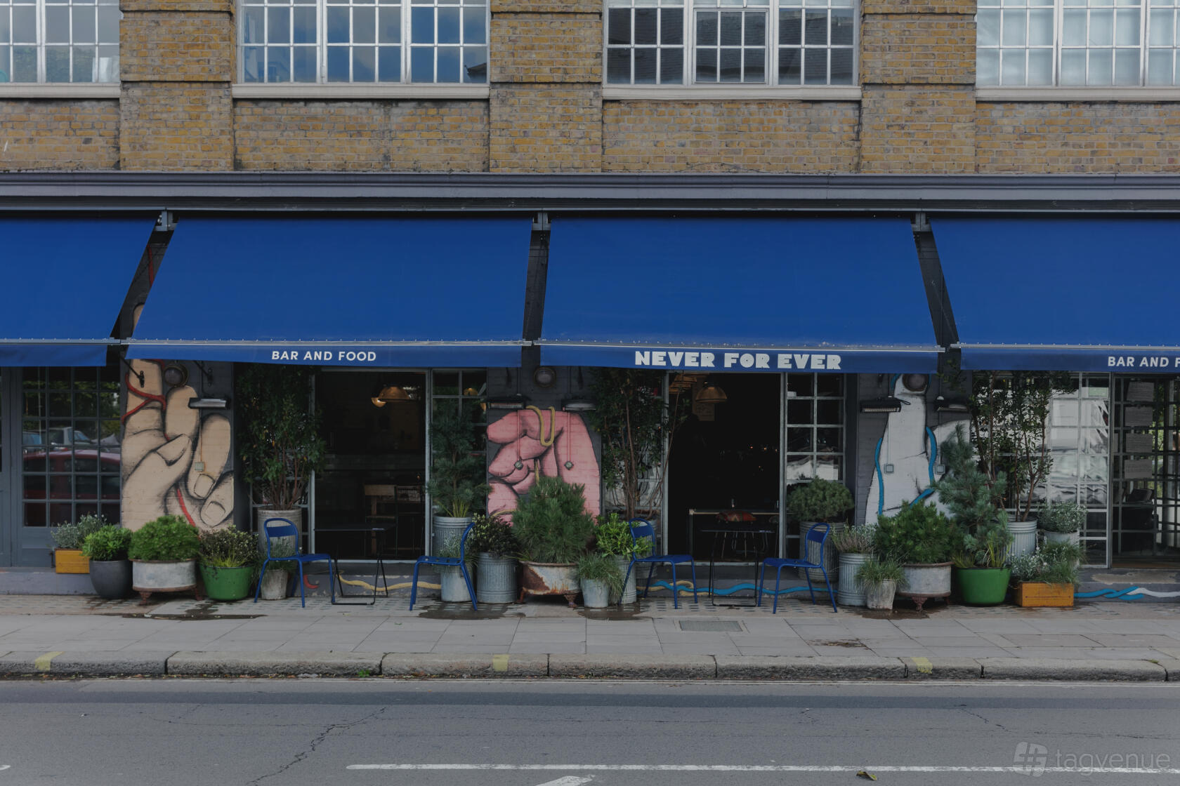 A pub with bold mural artwork, blue awning, and potted plants lining the entrance at Never For Ever.
