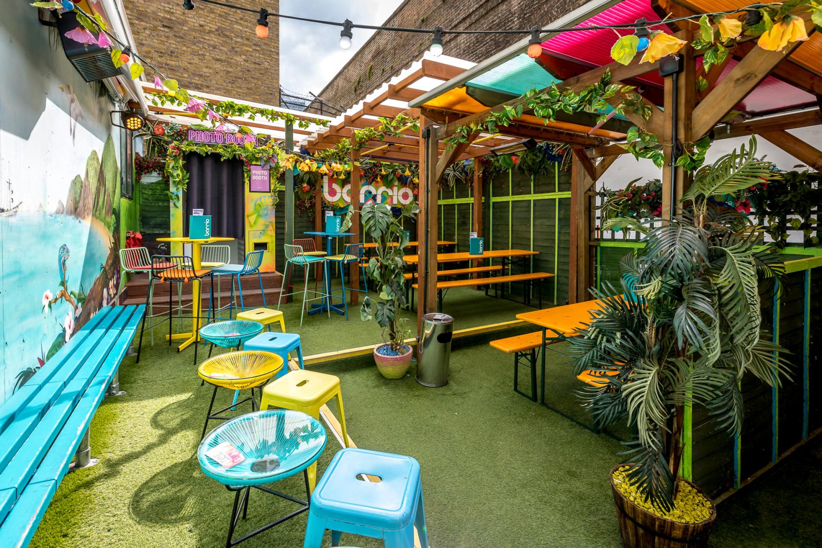 An outdoor bar area with artificial grass, colorful stools, wooden benches, and overhead string lights at Barrio Shoreditch.