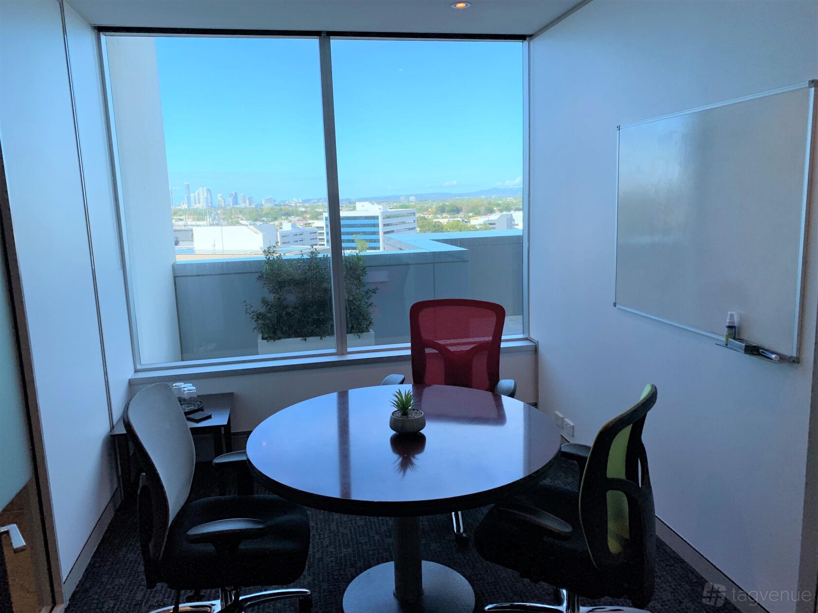 A meeting room with a round table, office chairs, large window with city view, and a wall-mounted whiteboard at @Workspaces - Bundall.