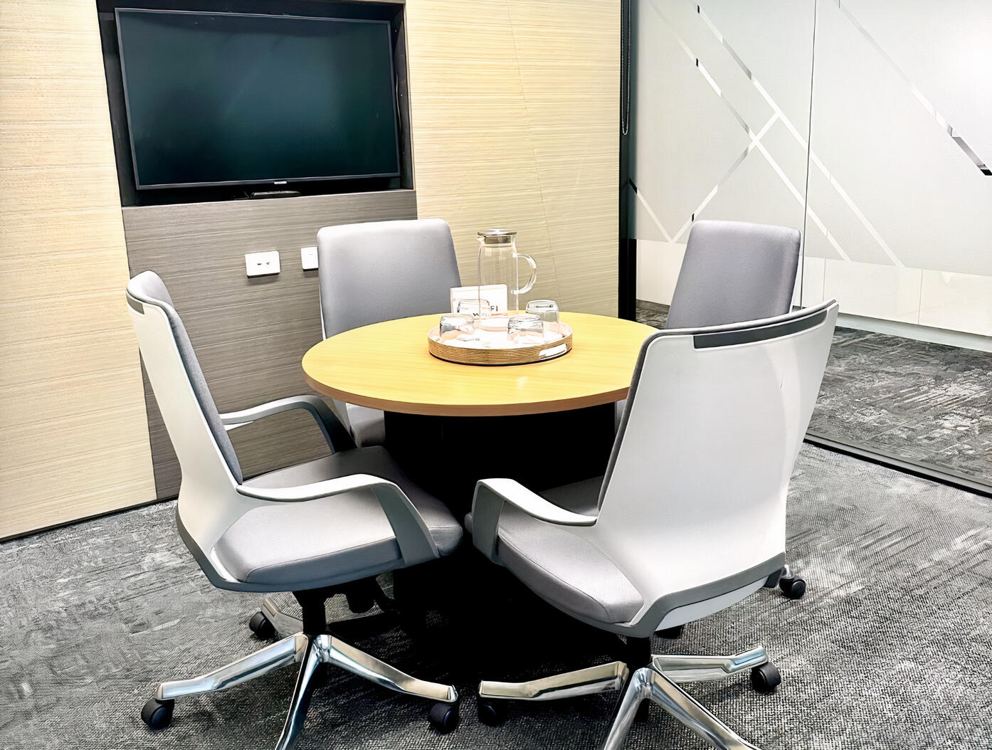 A meeting room with four grey office chairs, a round wooden table, and a wall-mounted TV at Compass Offices - 141 Walker Street.