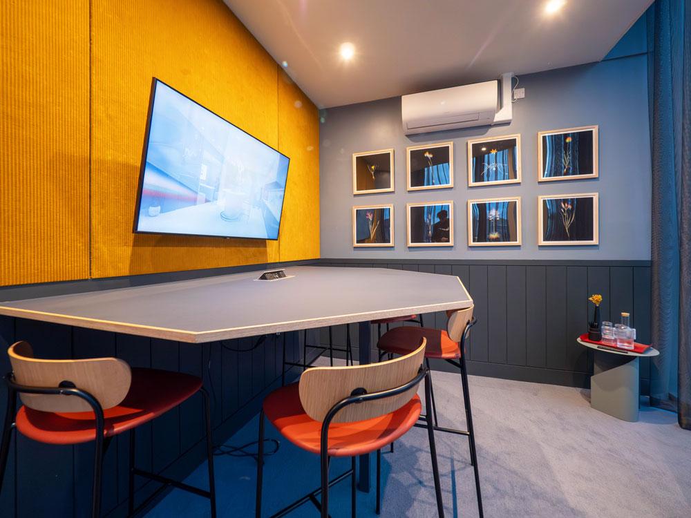 A meeting room with a wall-mounted screen, yellow accent wall, and four chairs at Colony Fabrica.