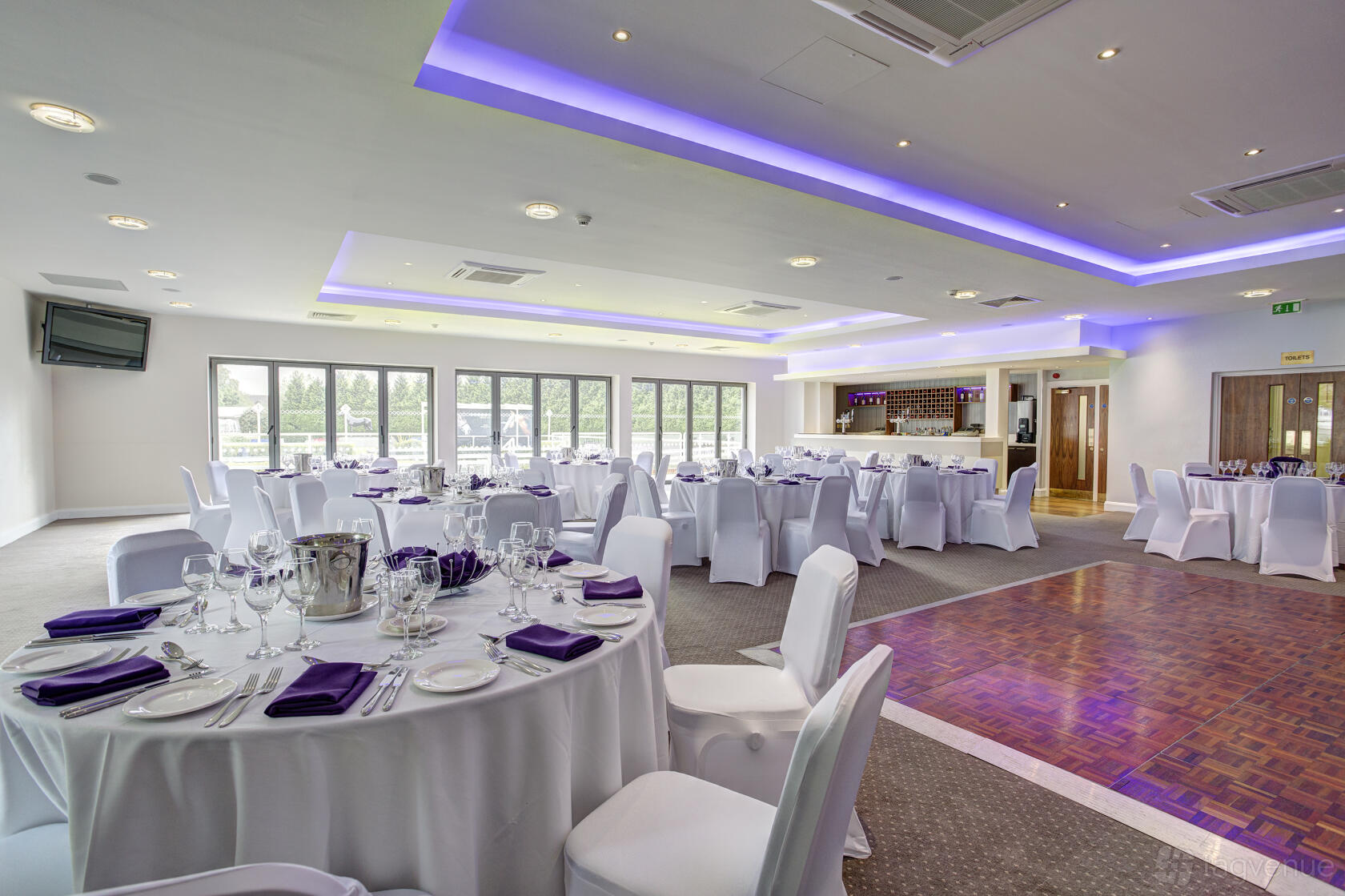 An event venue with round tables dressed in white linens, purple napkins, and a parquet dance floor at Kempton Park Racecourse.