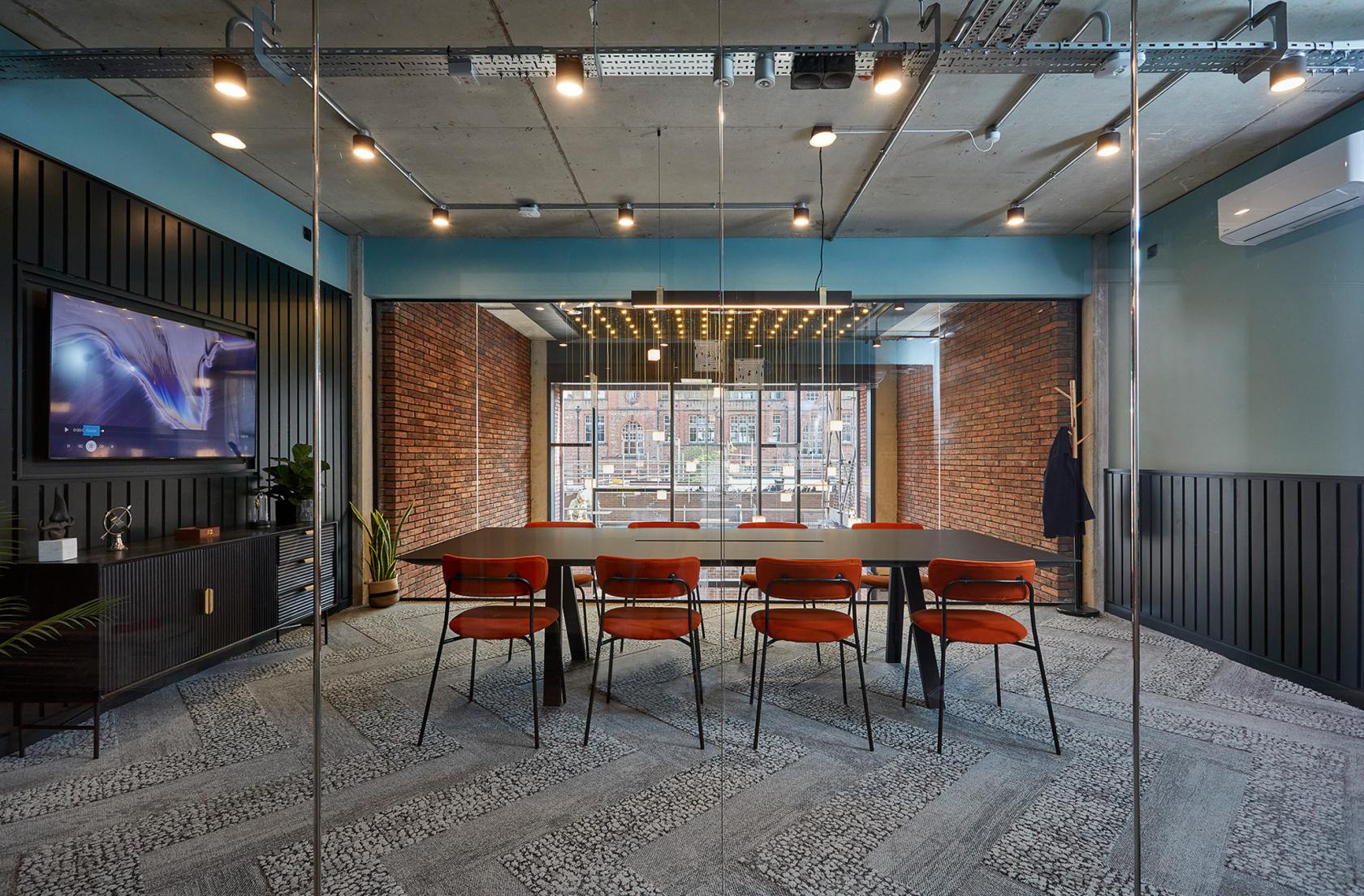 A meeting room with exposed brick walls, large windows, and a long table with orange chairs at Colony One Silk Street.