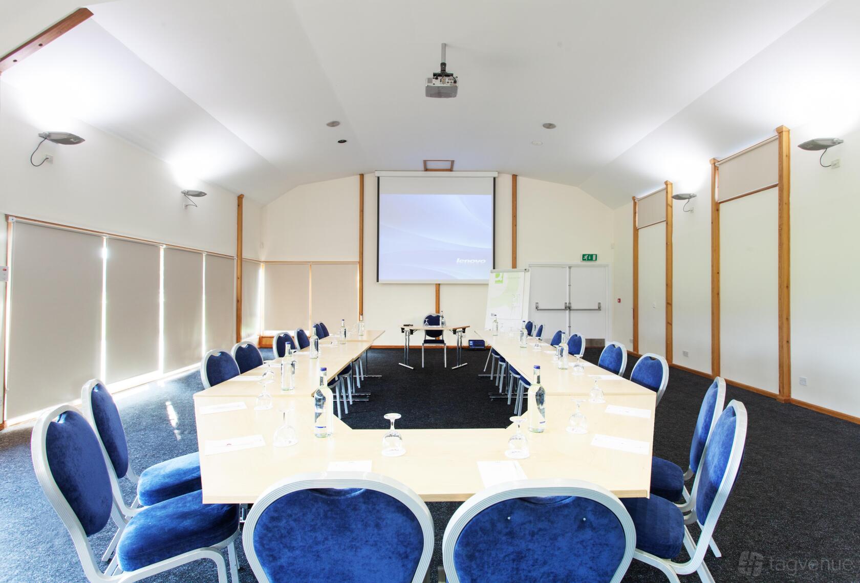 A meeting room with a U-shaped conference table, blue chairs, projector screen, and large windows at Kent Event Centre.