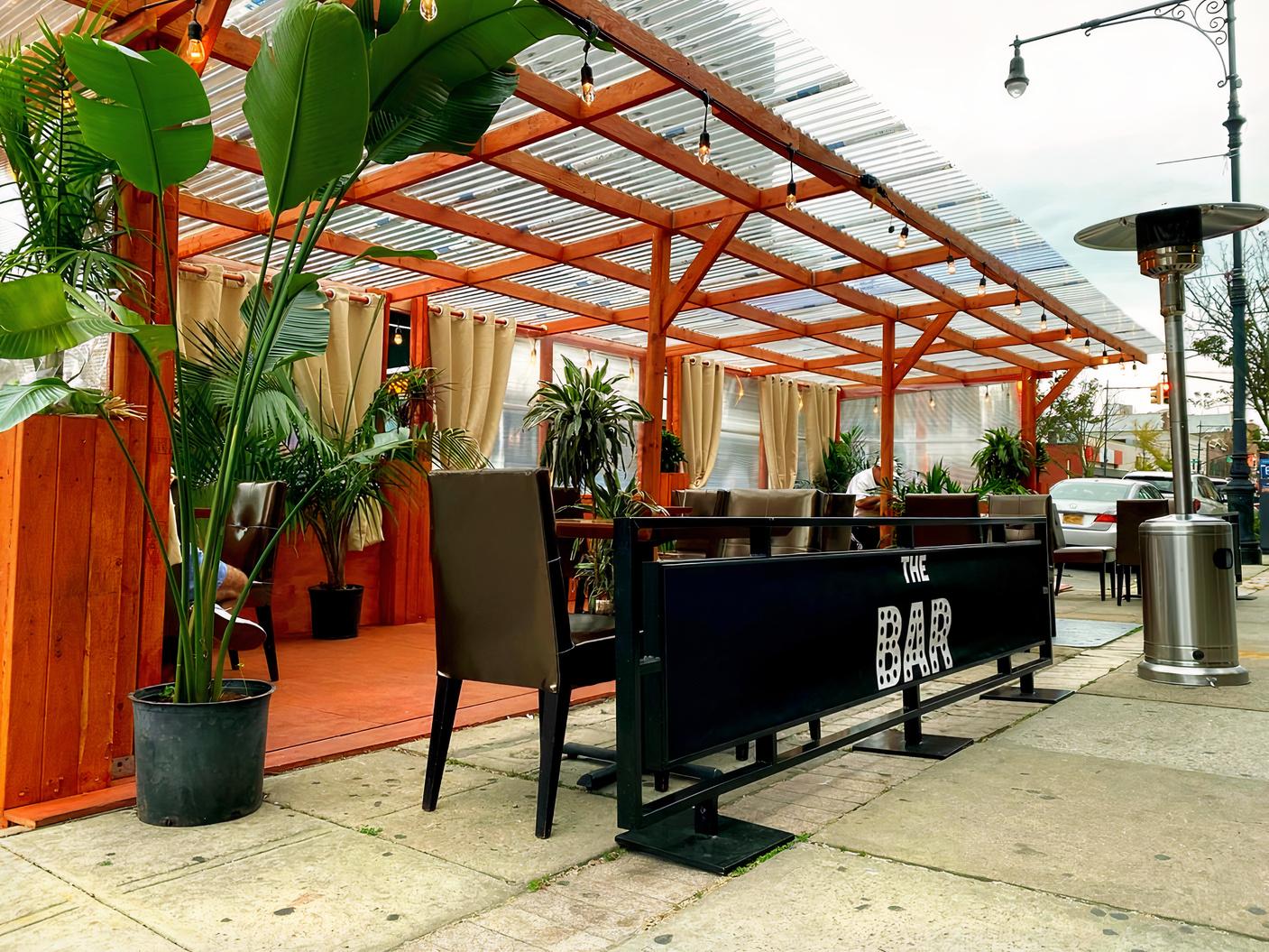An outdoor dining patio with wooden pergola, potted plants, and seating area at Zamaan Bay Ridge.