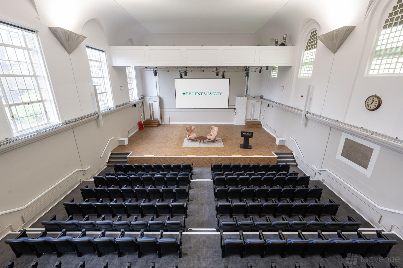 An auditorium with tiered black seating, high arched windows, and a stage with armchairs at Regent’s Events