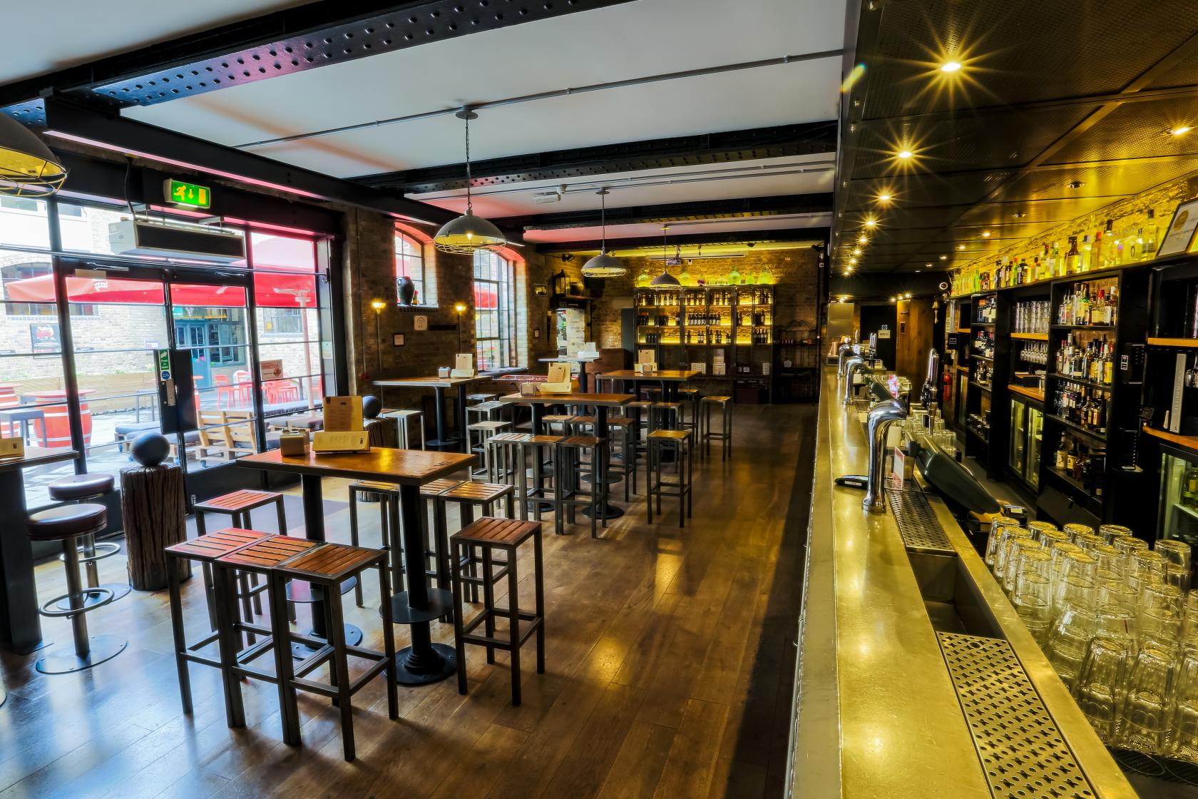 A bar in a restaurant with exposed brick walls, high tables, wooden flooring, and large windows at Camino King's Cross.