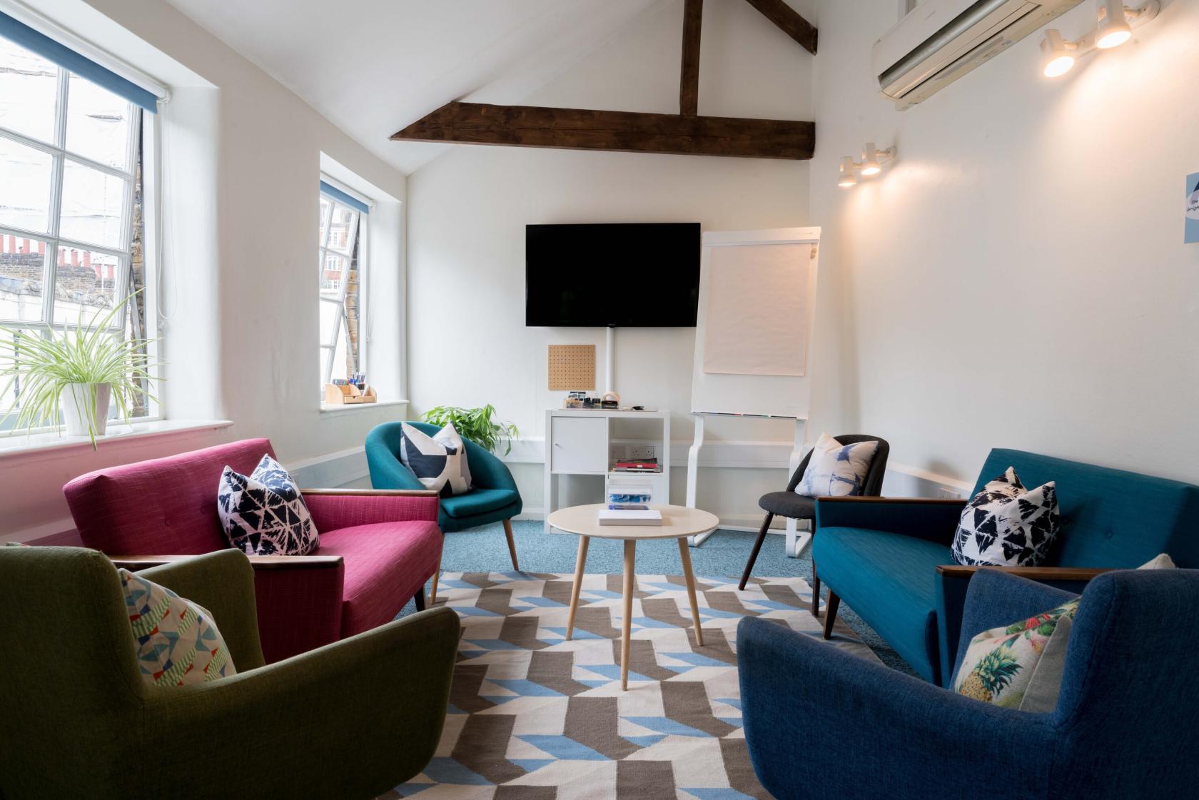 An event venue room with colorful armchairs, patterned rug, wall-mounted TV, and large windows at Wallacespace St Pancras.