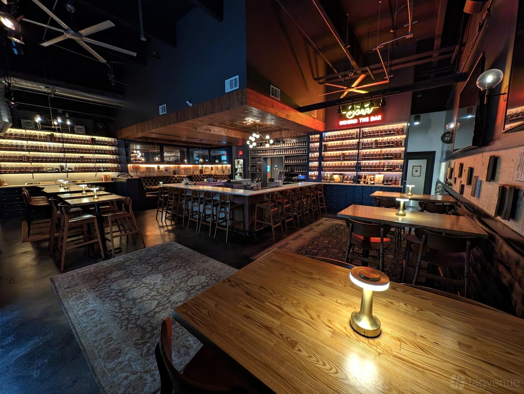 An event space in a restaurant with shelves of whiskey bottles, wooden tables, and glowing lamps at Standard Proof Whiskey.