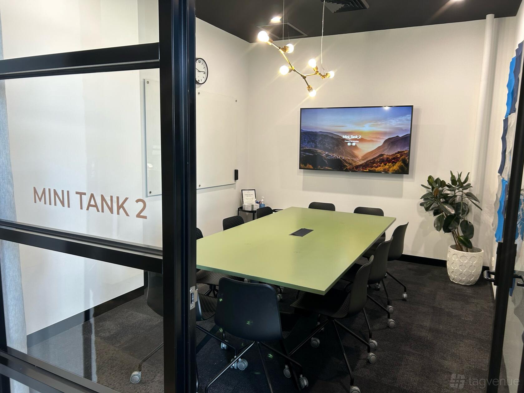A meeting room with a green conference table, black chairs, wall-mounted screen, and whiteboard at Work Tank Coworking & Serviced Offices.