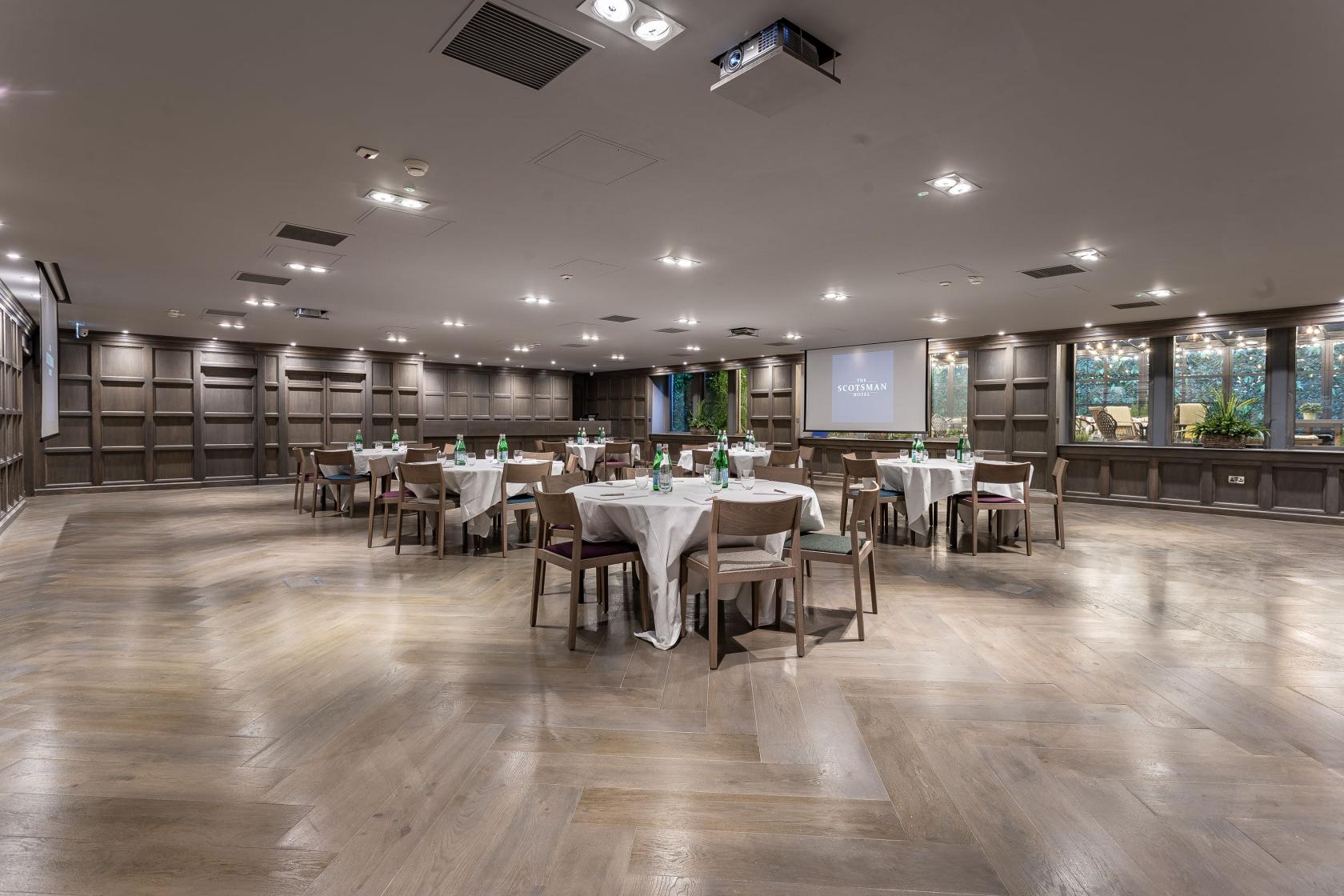 A function room with wood panelled walls, round tables with white linens, and a projector screen at The Scotsman Hotel.
