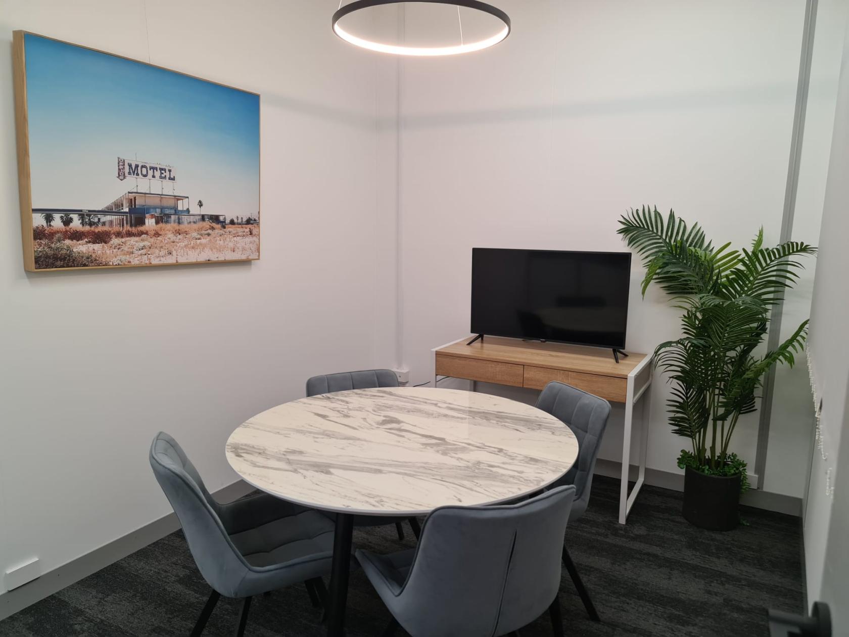 A meeting room with a round marble table, four gray chairs, wall art, a TV, and a potted plant at Workplex.