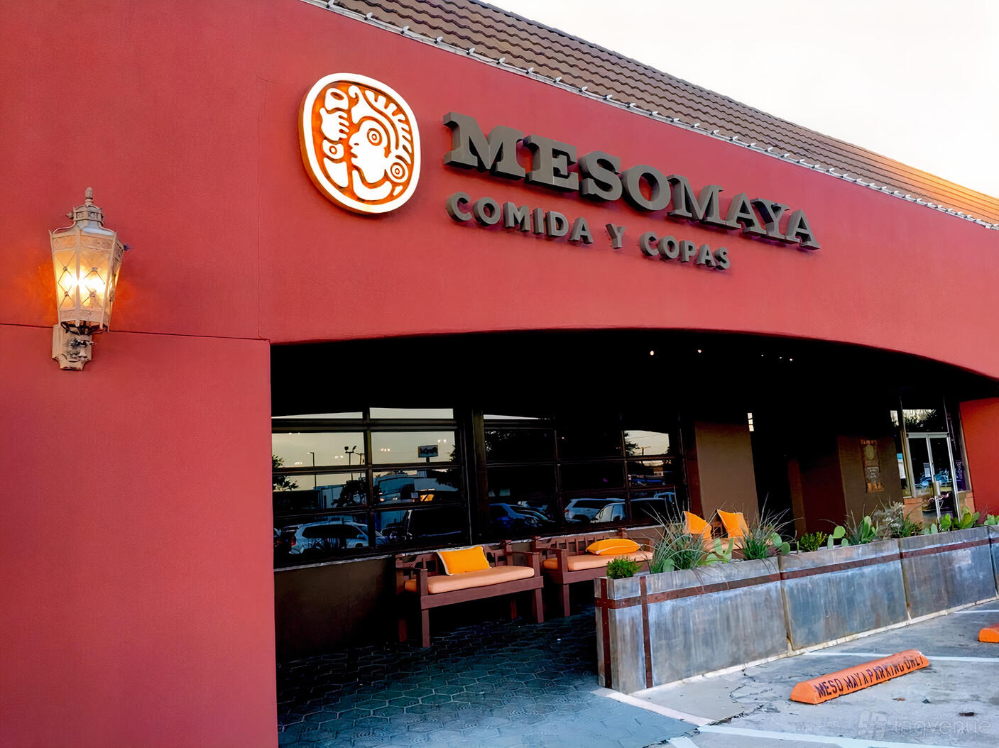 A restaurant exterior with a red stucco facade, orange signage, and outdoor bench seating at Meso Maya - Preston Forest.