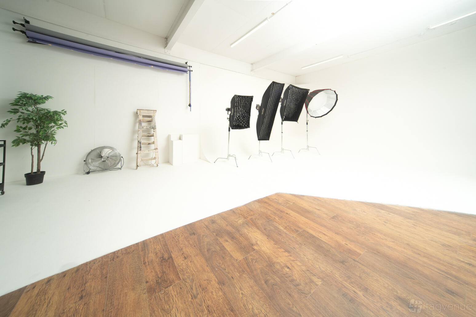A photography studio with seamless white walls, wooden flooring, large softbox lights, and backdrop rolls at Phos Media studios.