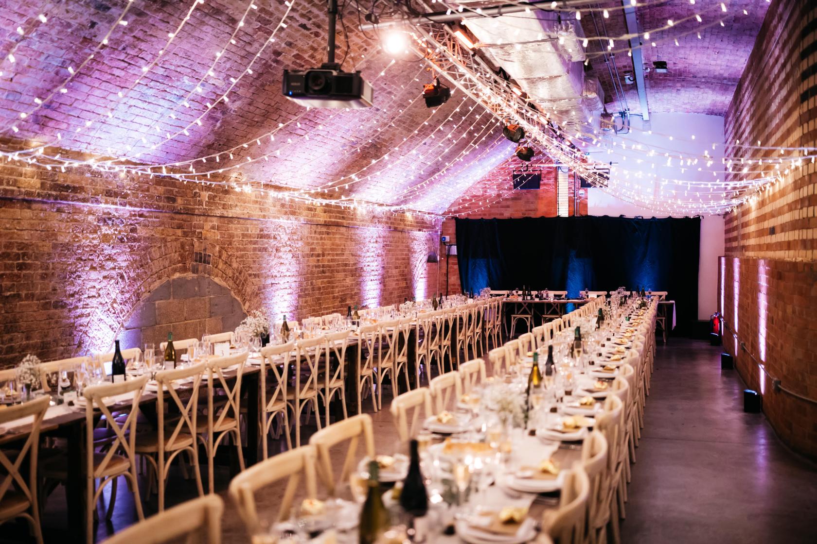 A warehouse event space with exposed brick arches, long banquet tables, and string lighting at Kachette.
