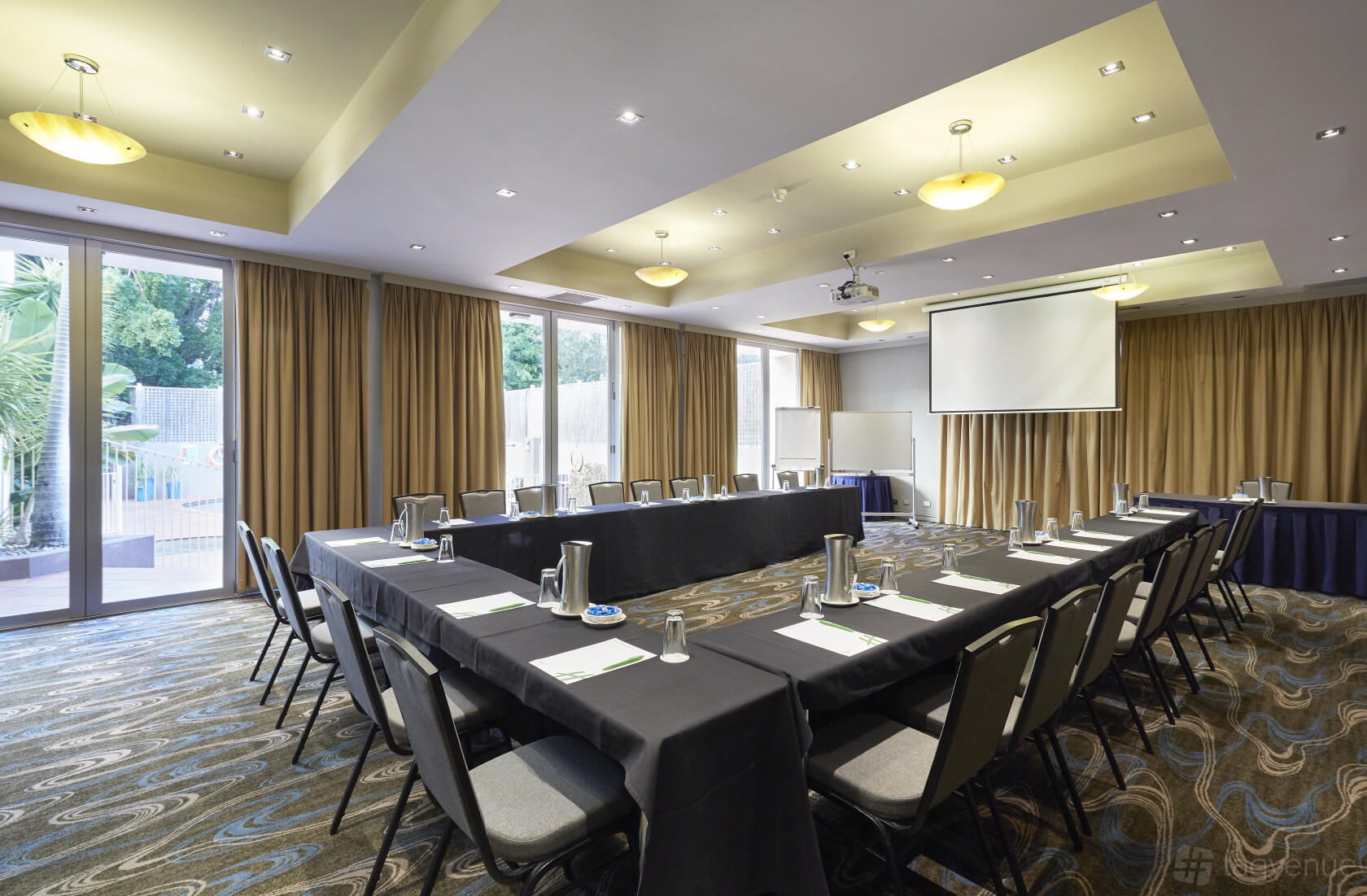A hotel meeting room with U-shaped tables, floor-to-ceiling windows, brown curtains, and a ceiling projector at Holiday Inn Parramatta.