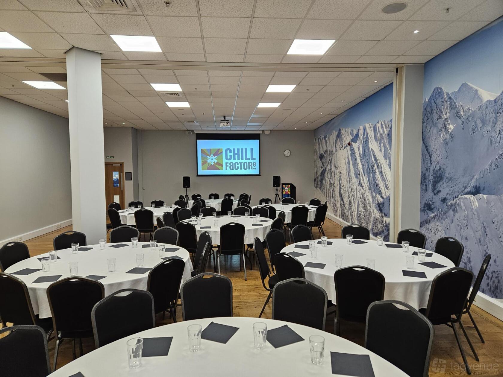 An event space with round tables, black chairs, a presentation screen, and a mountain mural at Chill Factore.
