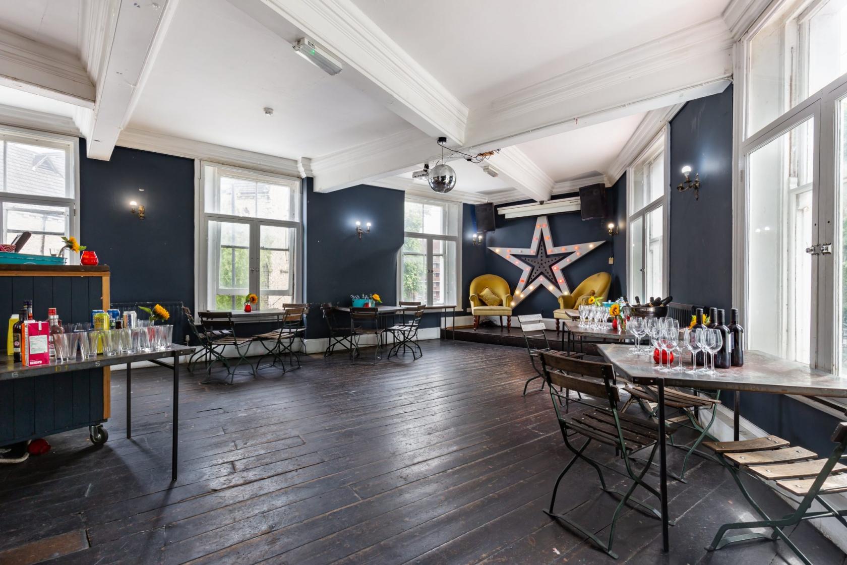 A function room with dark wood floors, large windows, metal chairs, and a stage with a star backdrop at Star by Hackney Downs.