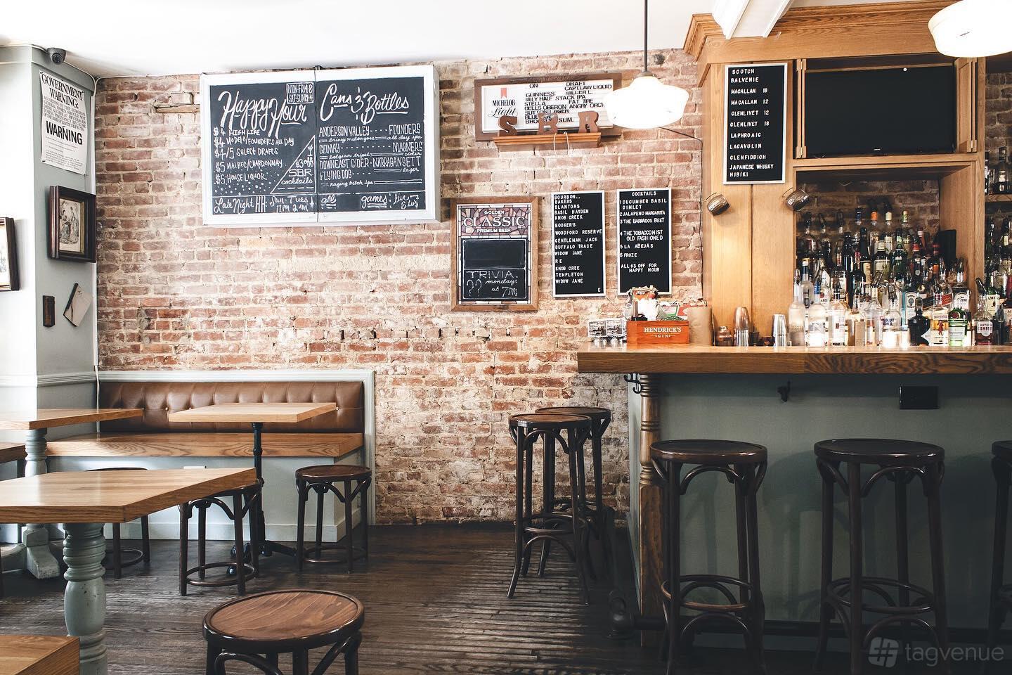 An event space in a bar with exposed brick walls, a stocked bar counter, and wooden tables at Sutton Bar Room.