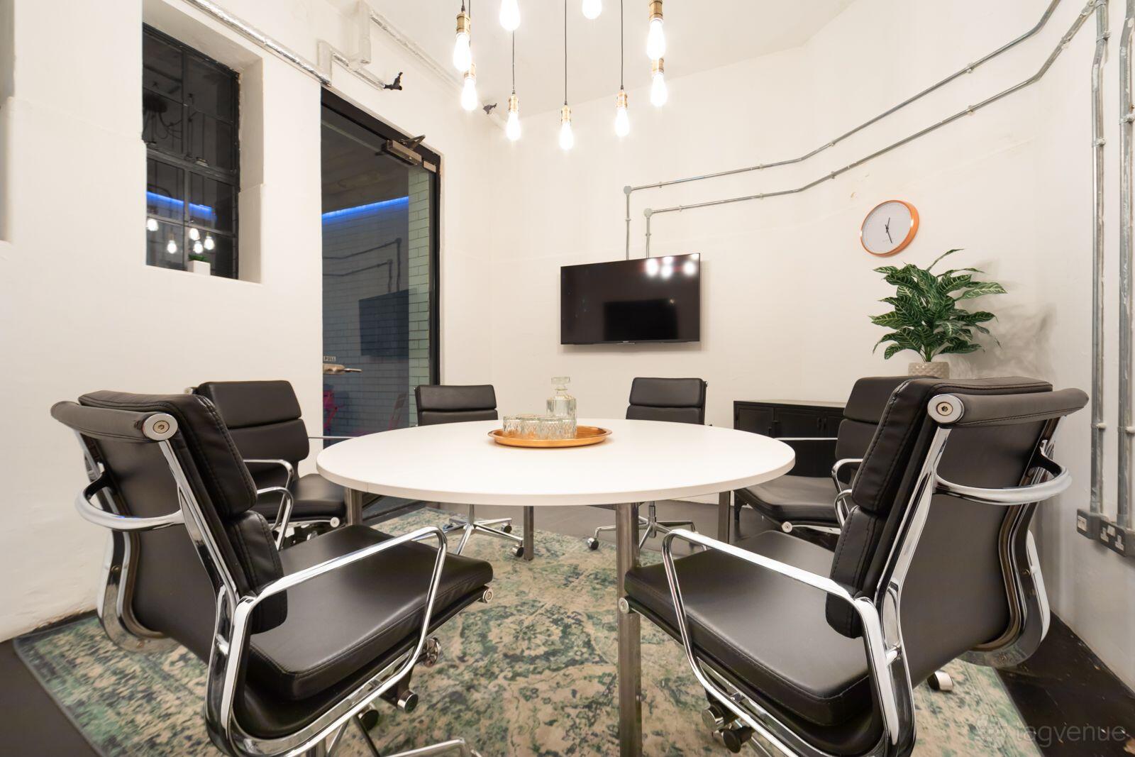A meeting room with a round table, six black chairs, wall-mounted TV, hanging lights, and a plant at The Space Holborn.