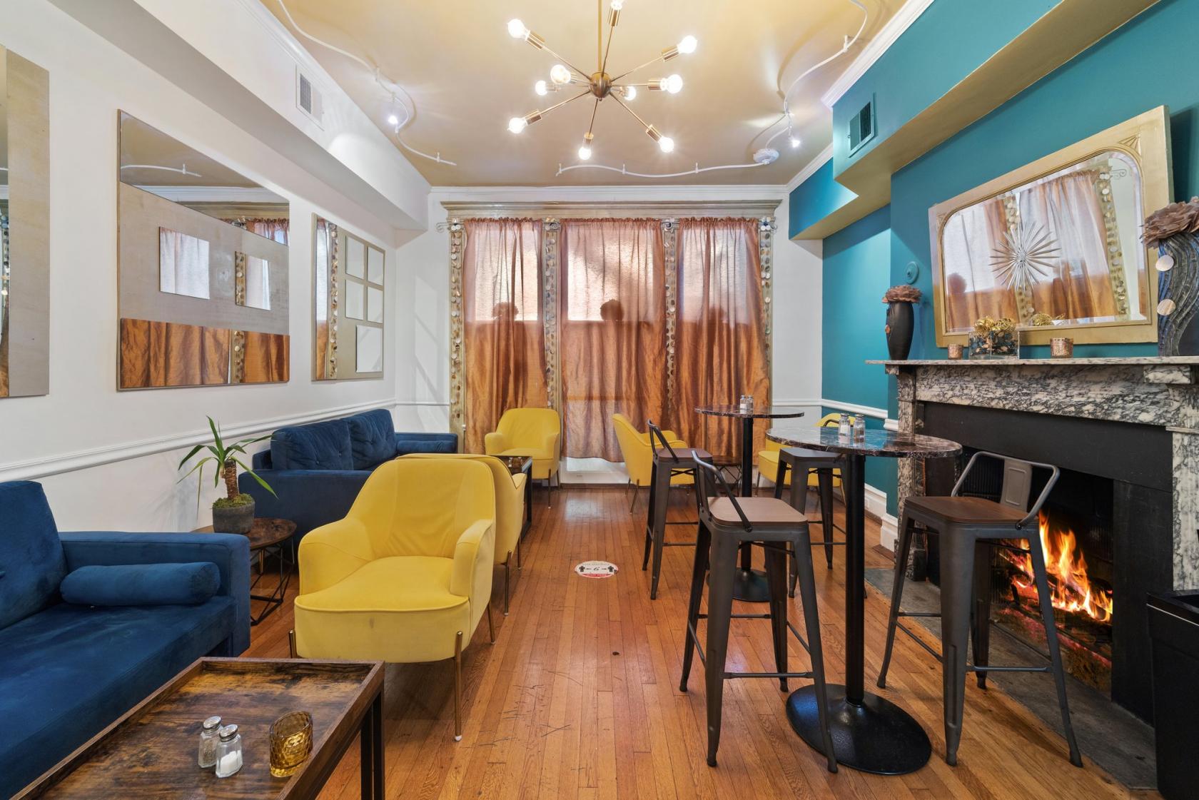 A restaurant with yellow and blue lounge chairs, small round tables, and a lit fireplace at Petit Monde Cafe.