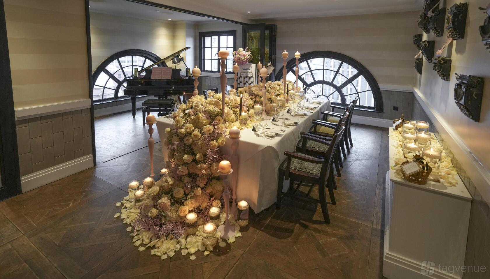 A hotel dining room with arched windows, a grand piano, and a banquet table decorated with florals and candles at Hotel Gotham.