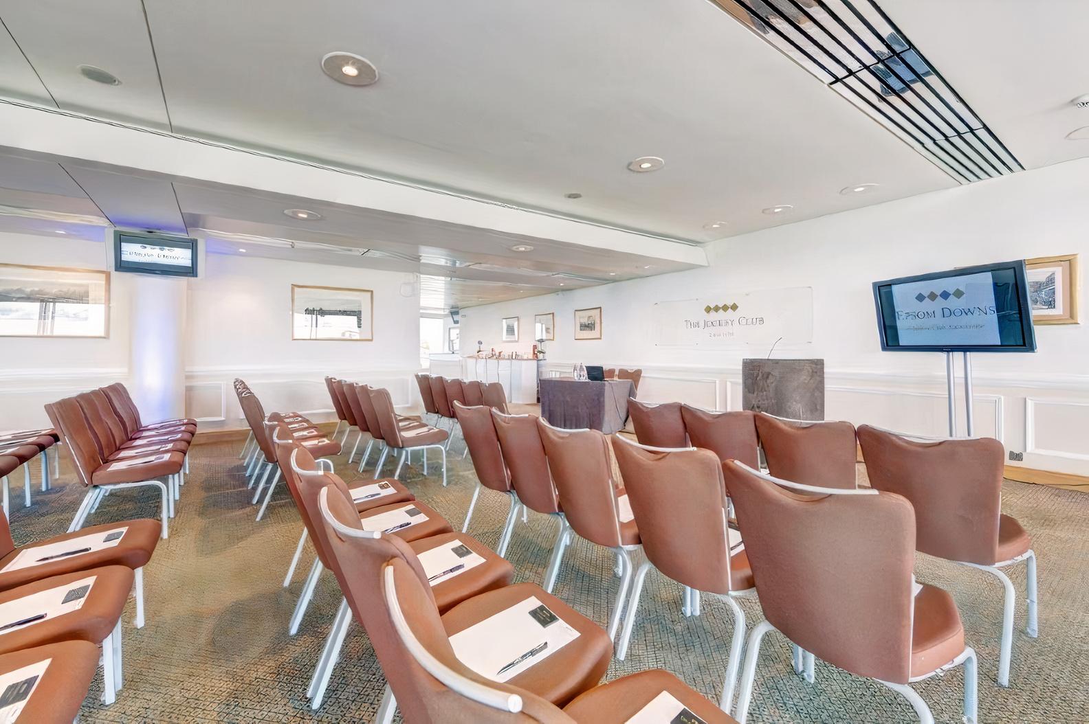 An event venue with rows of brown chairs facing a presentation area and large windows at Epsom Downs Racecourse.