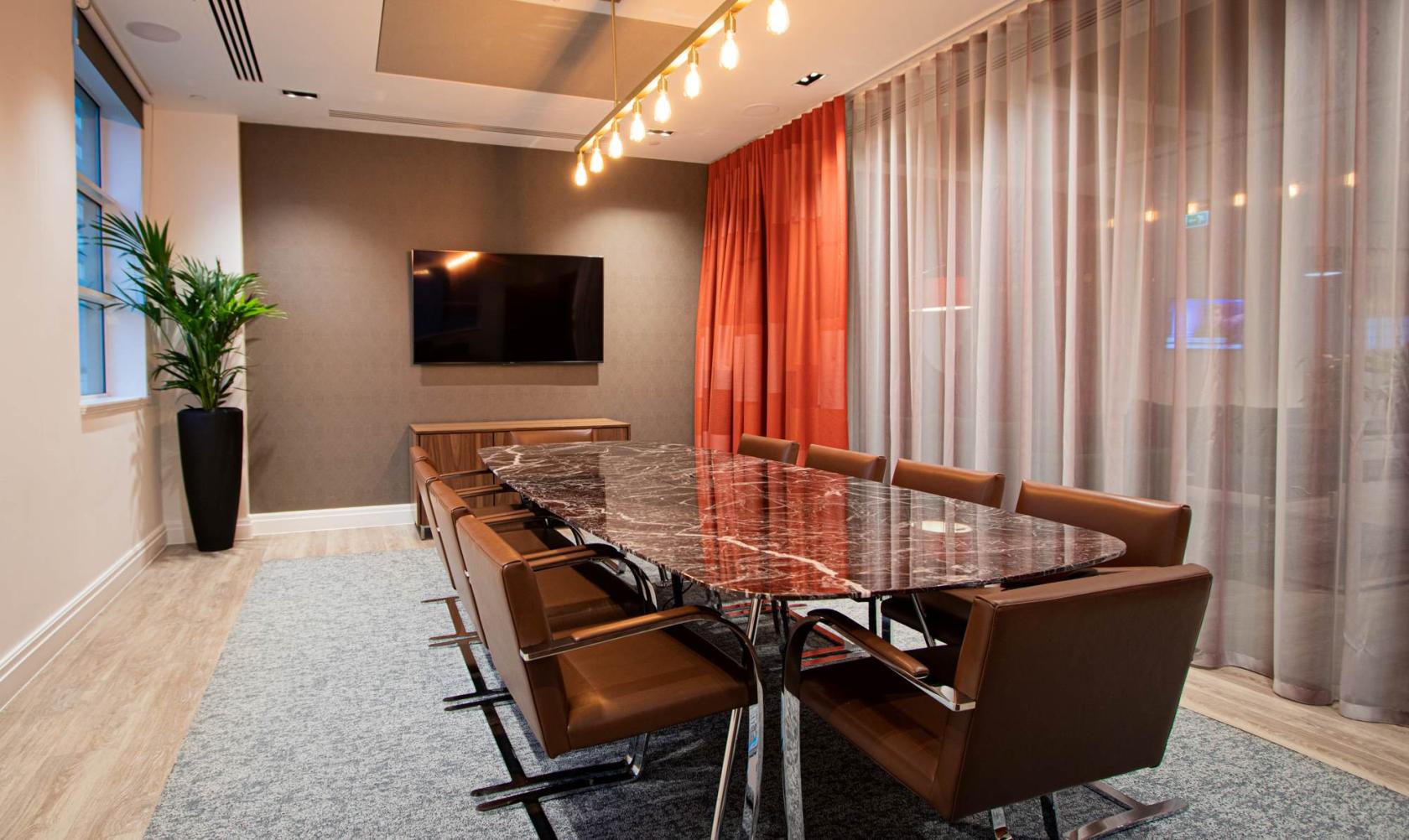 A meeting room with a marble conference table, brown leather chairs, wall-mounted TV, and sheer curtains at Gilbanks, One Park Row.