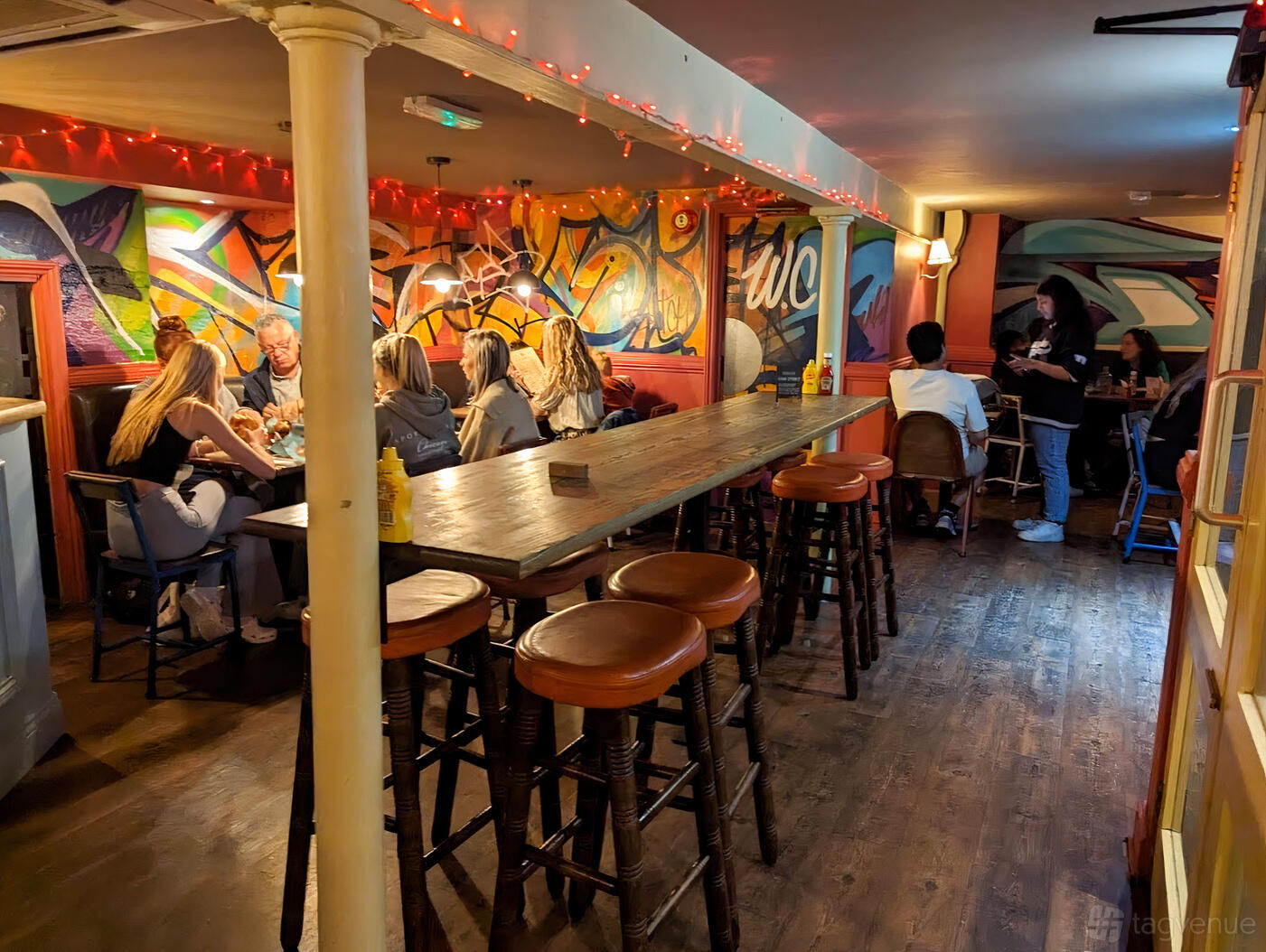 A restaurant with colorful graffiti-style wall art, wooden floors, and high tables with stools at Butch Annie’s Restaurant.