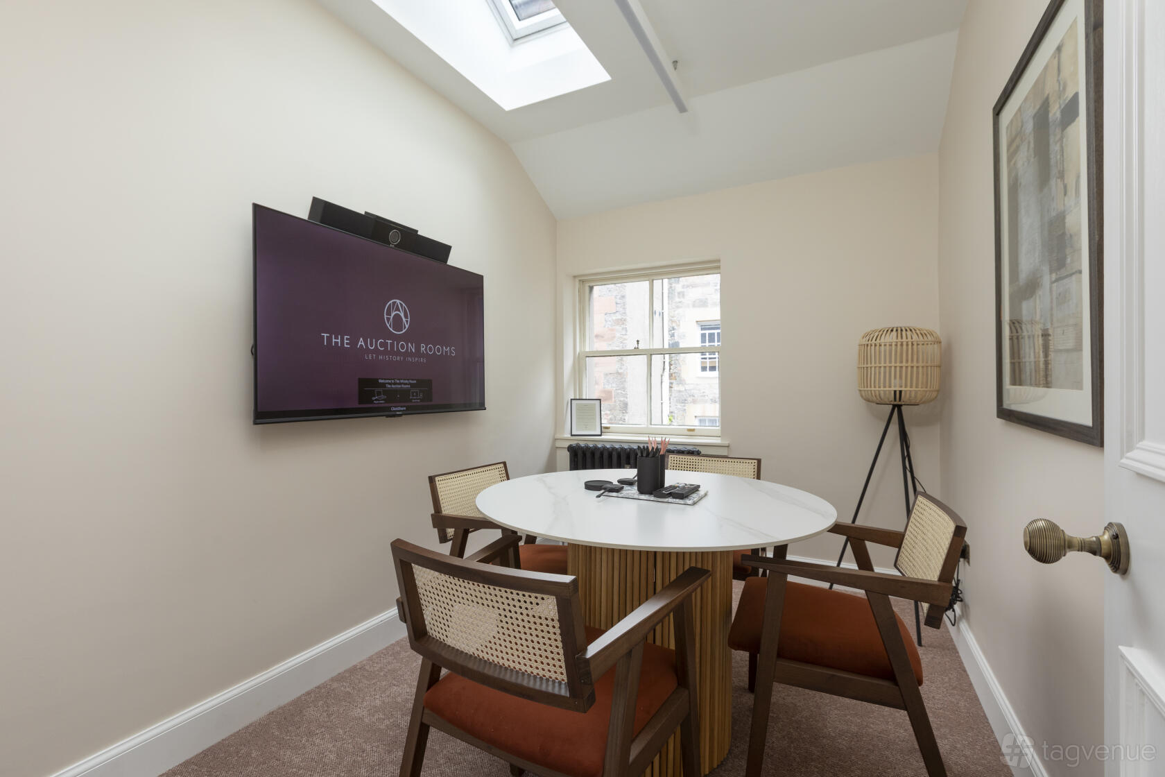 A meeting room with a round white table, four cushioned chairs, wall-mounted screen, and skylight at The Auction Rooms.