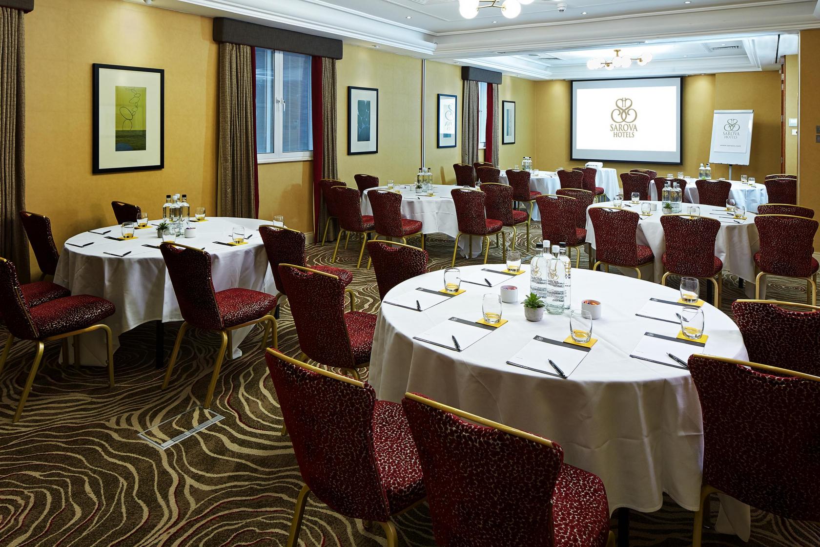 A hotel function room with round tables, red upholstered chairs, and a projector screen at The Rembrandt Hotel.