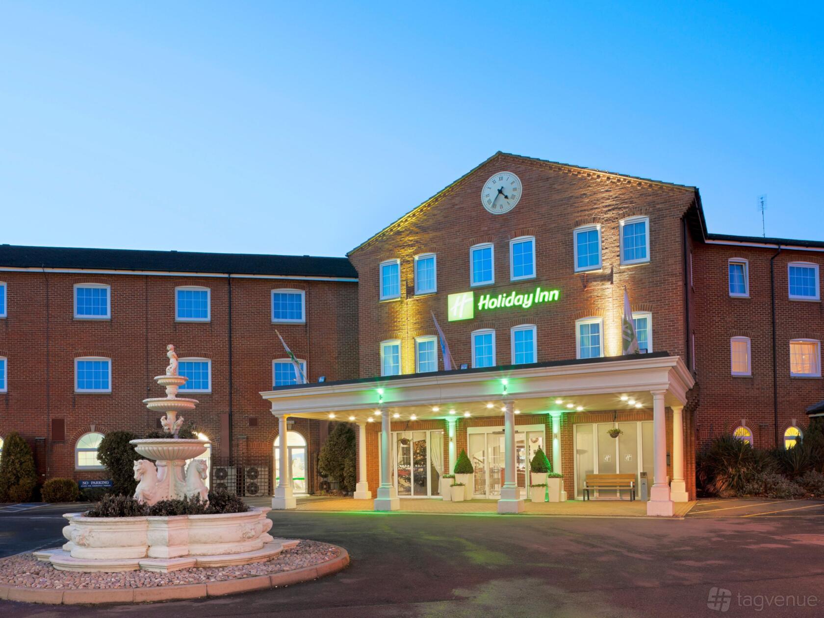 A hotel with a brick exterior, white columned entrance, illuminated sign, and a tiered fountain at Holiday Inn Corby.