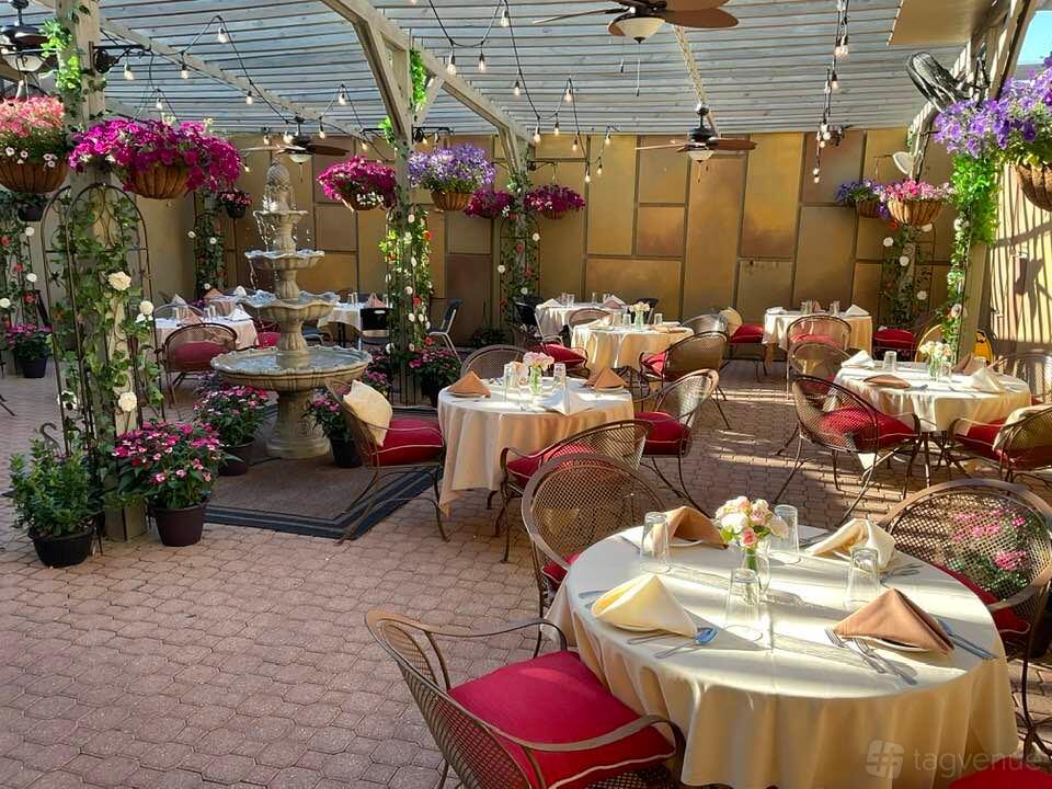 A restaurant terrace with round tables, wicker chairs, hanging flower baskets, and a central fountain at Bella Mia Fine Dining.