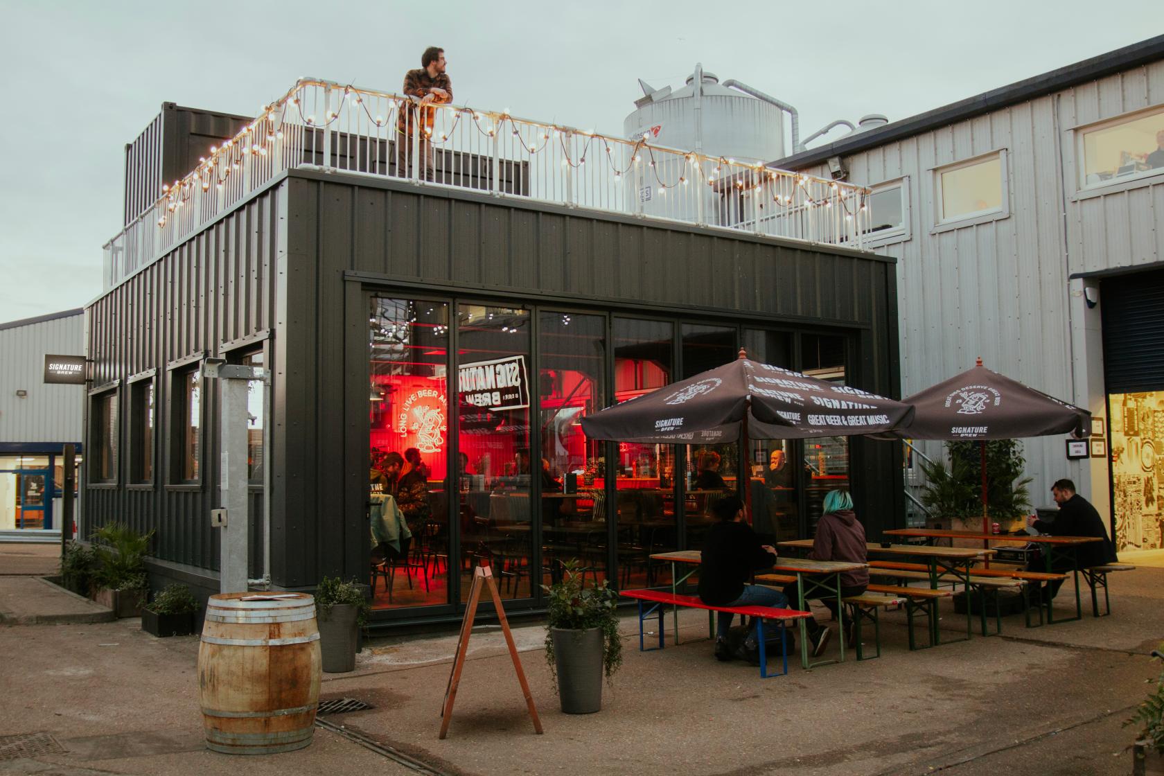 A terrace at a brewery with festoon lights, outdoor picnic tables, and branded umbrellas at Signature Brew Blackhorse Road.
