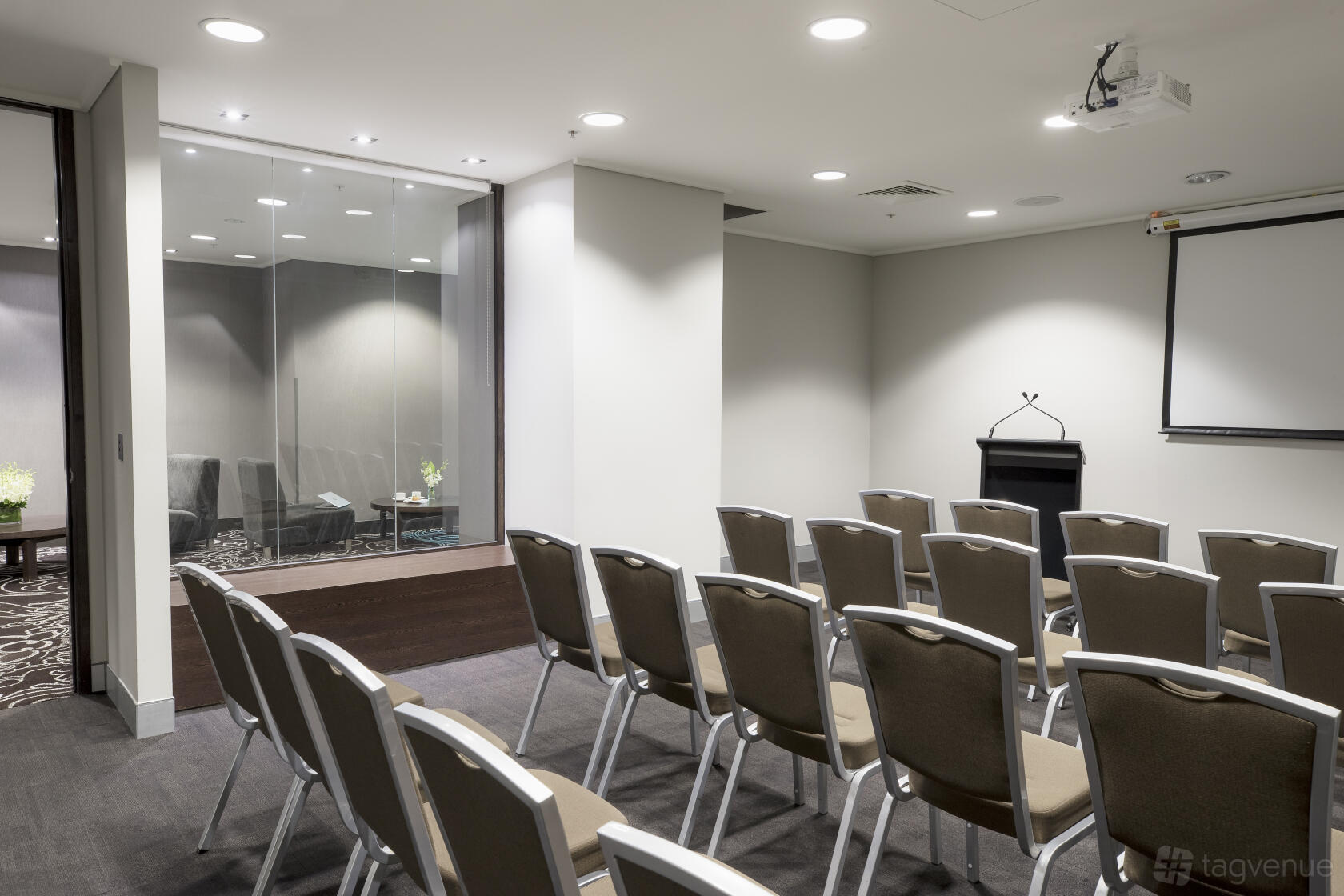 A hotel meeting room with rows of beige chairs, a podium, projector, and glass partition at Novotel Melbourne on Collins.