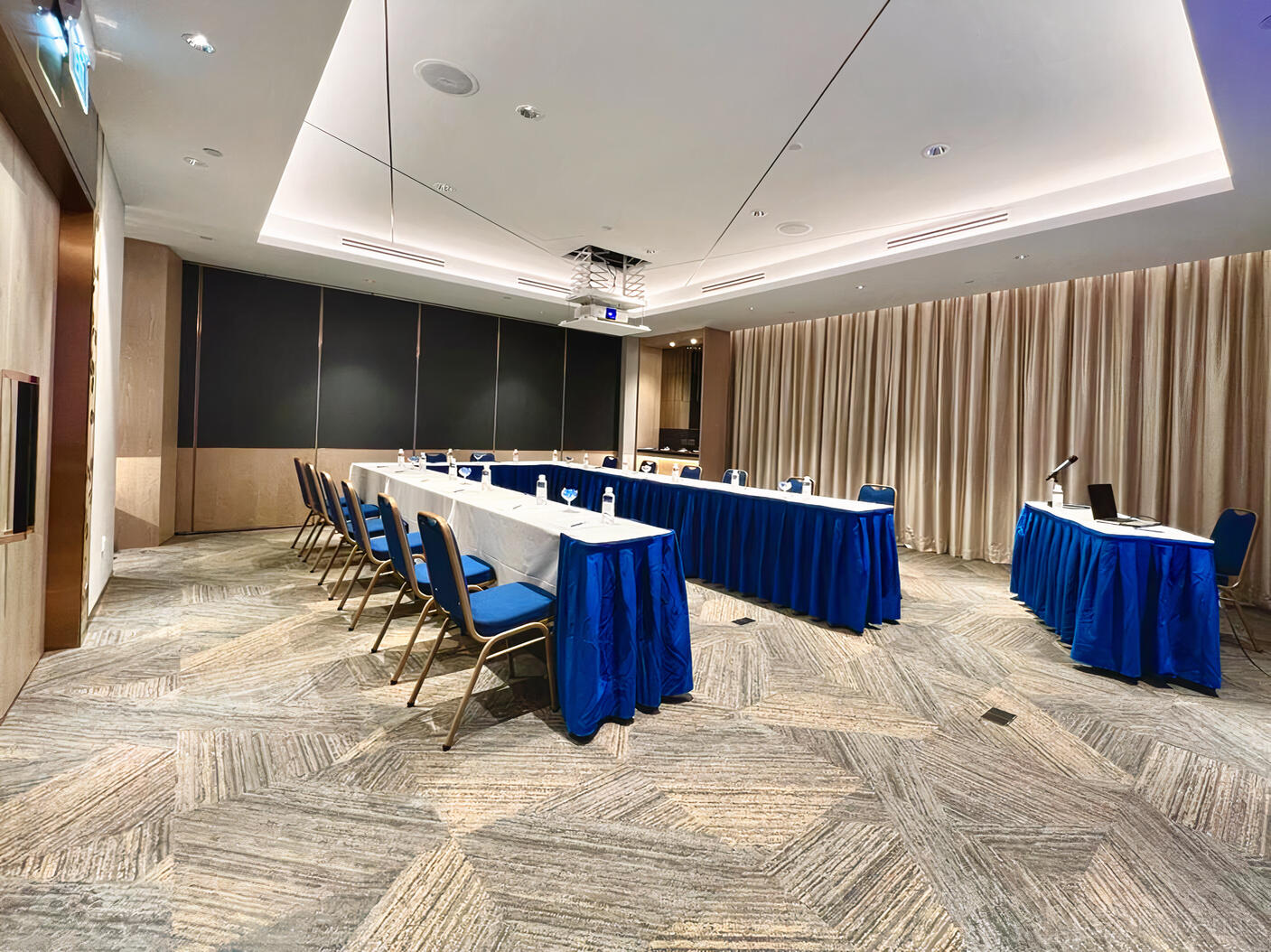 A hotel meeting room with U-shaped tables with blue skirting, presentation screen, and carpet flooring at Orchard Grand Court.