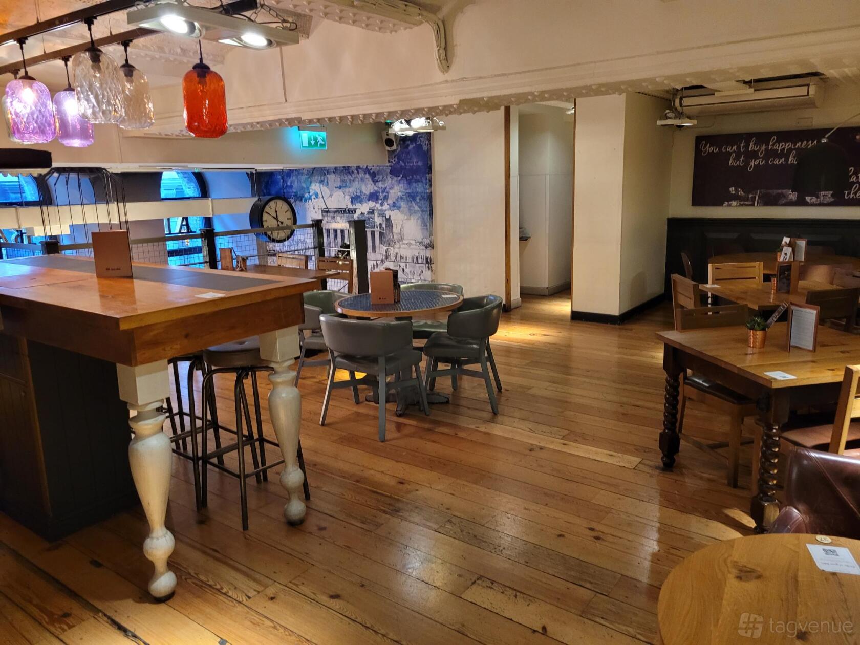 An event space in a bar with wooden floors, round tables, colorful pendant lights, and wall art at All Bar One Charing Cross.