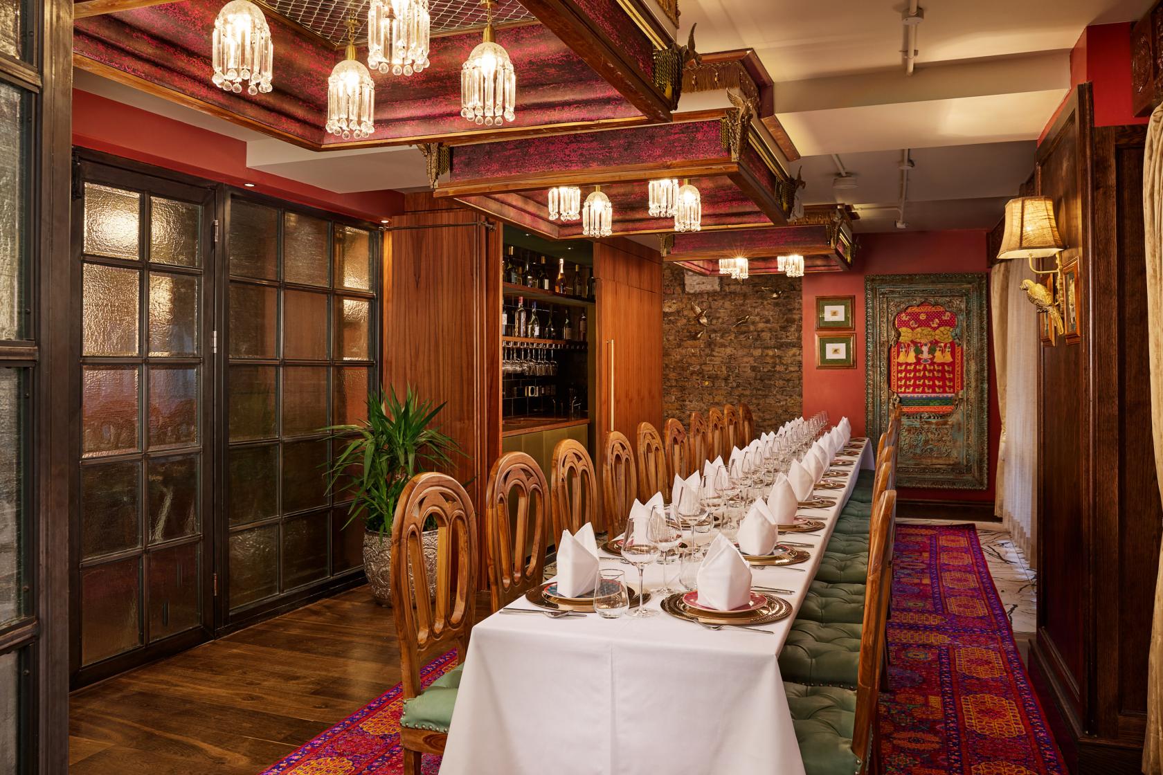 A dining room with a long table set with white linens, ornate wooden chairs, and decorative ceiling lights at Colonel Saab Trafalgar Square.