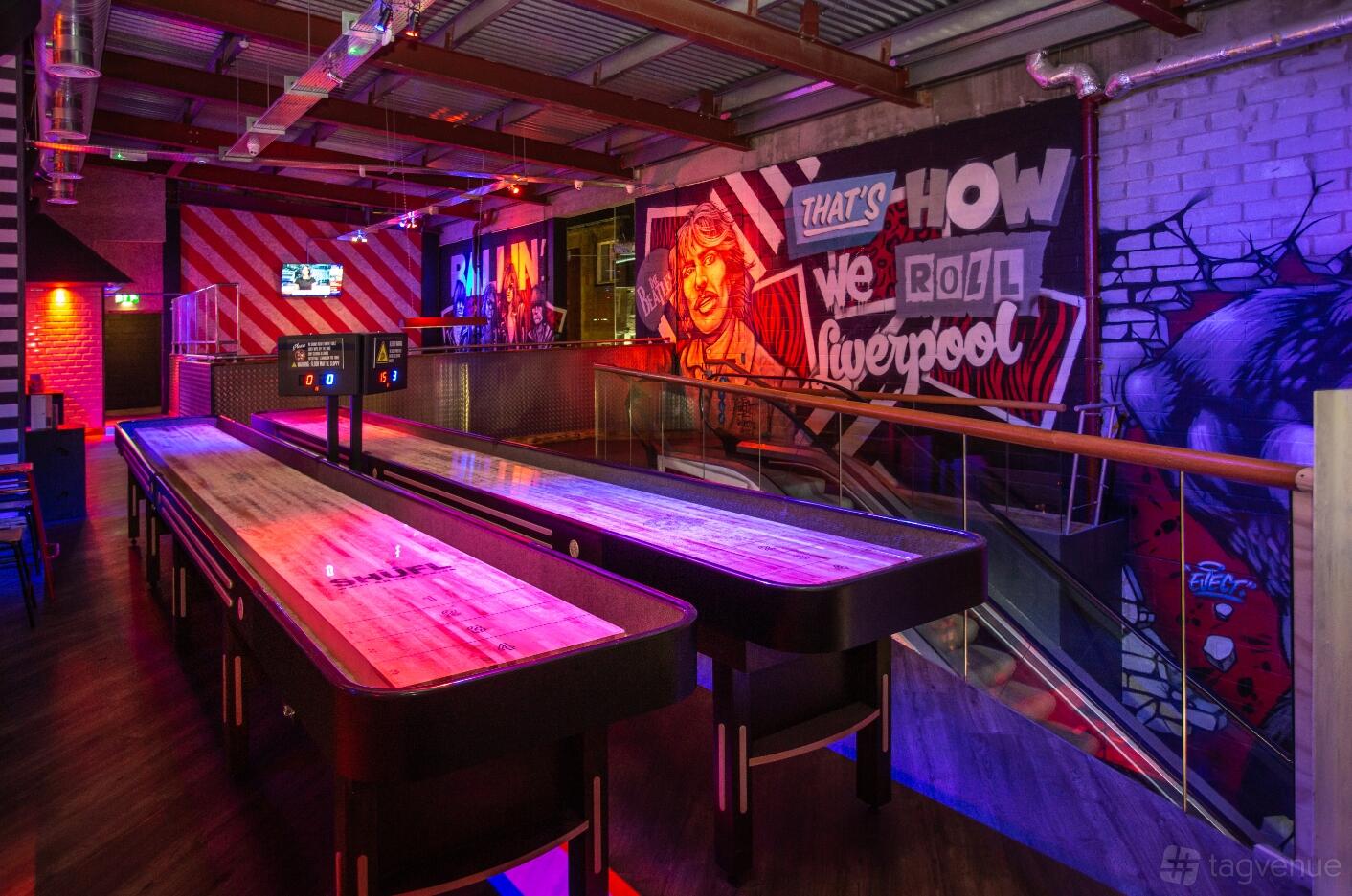 A bar with shuffleboard tables, exposed ductwork, colorful graffiti walls, and neon lighting at Roxy Ball Room Liverpool Cavern Quarter.