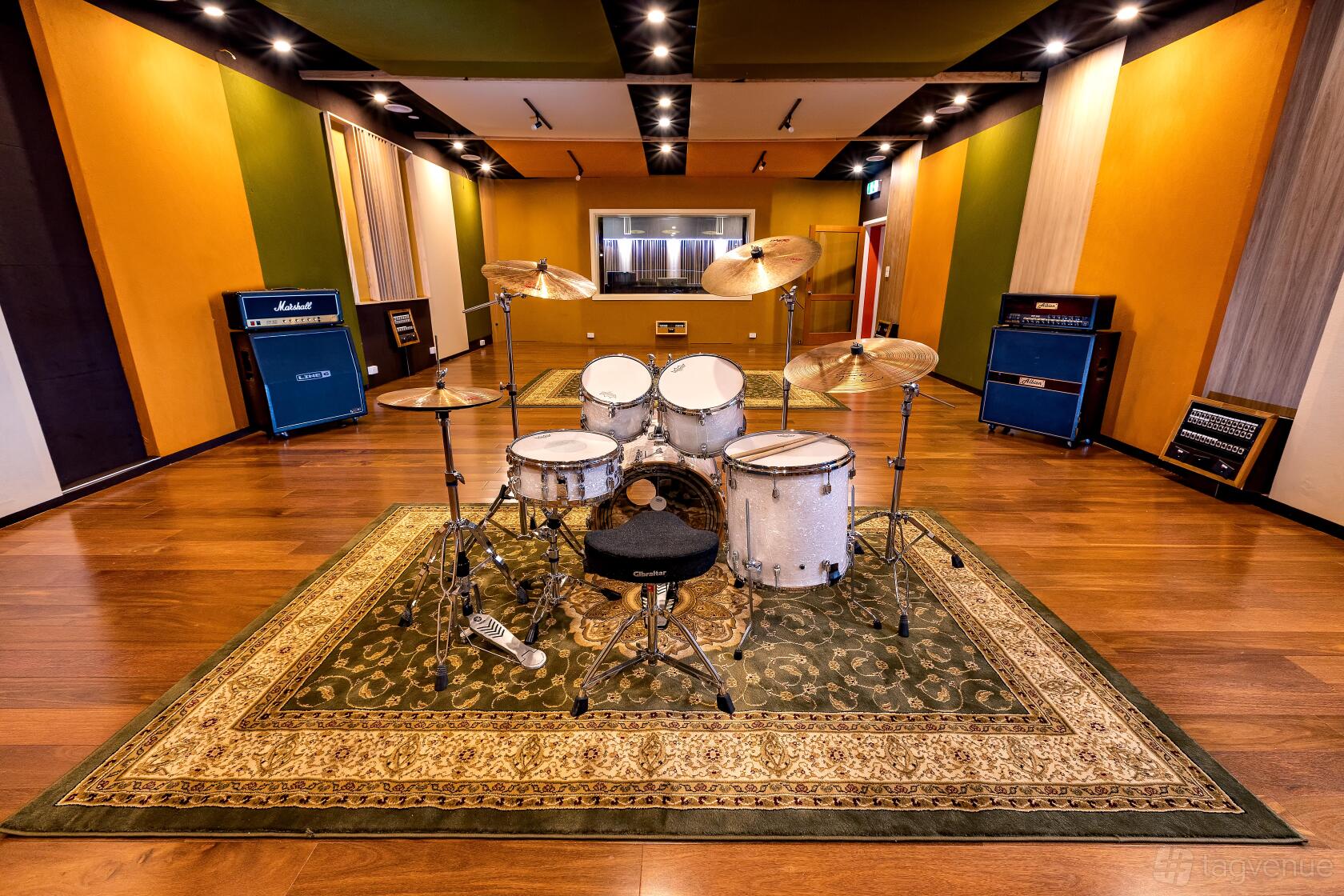 A rehearsal room with a drum set on a patterned rug, wood floors, amplifiers, and soundproofed walls at Empire Music Studios A.K.A Studio52.