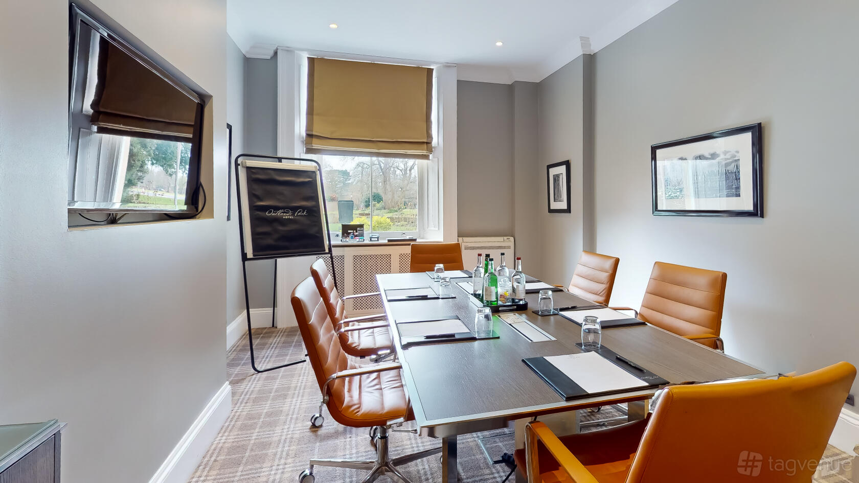 A boardroom with a rectangular table, tan leather chairs, flip chart, and large window at Oatlands Park Hotel.