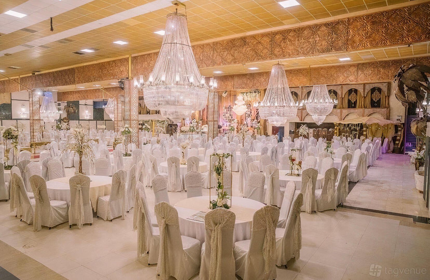 An event venue with crystal chandeliers, gold accents, and round tables draped in white linens at Excellency Bolton.