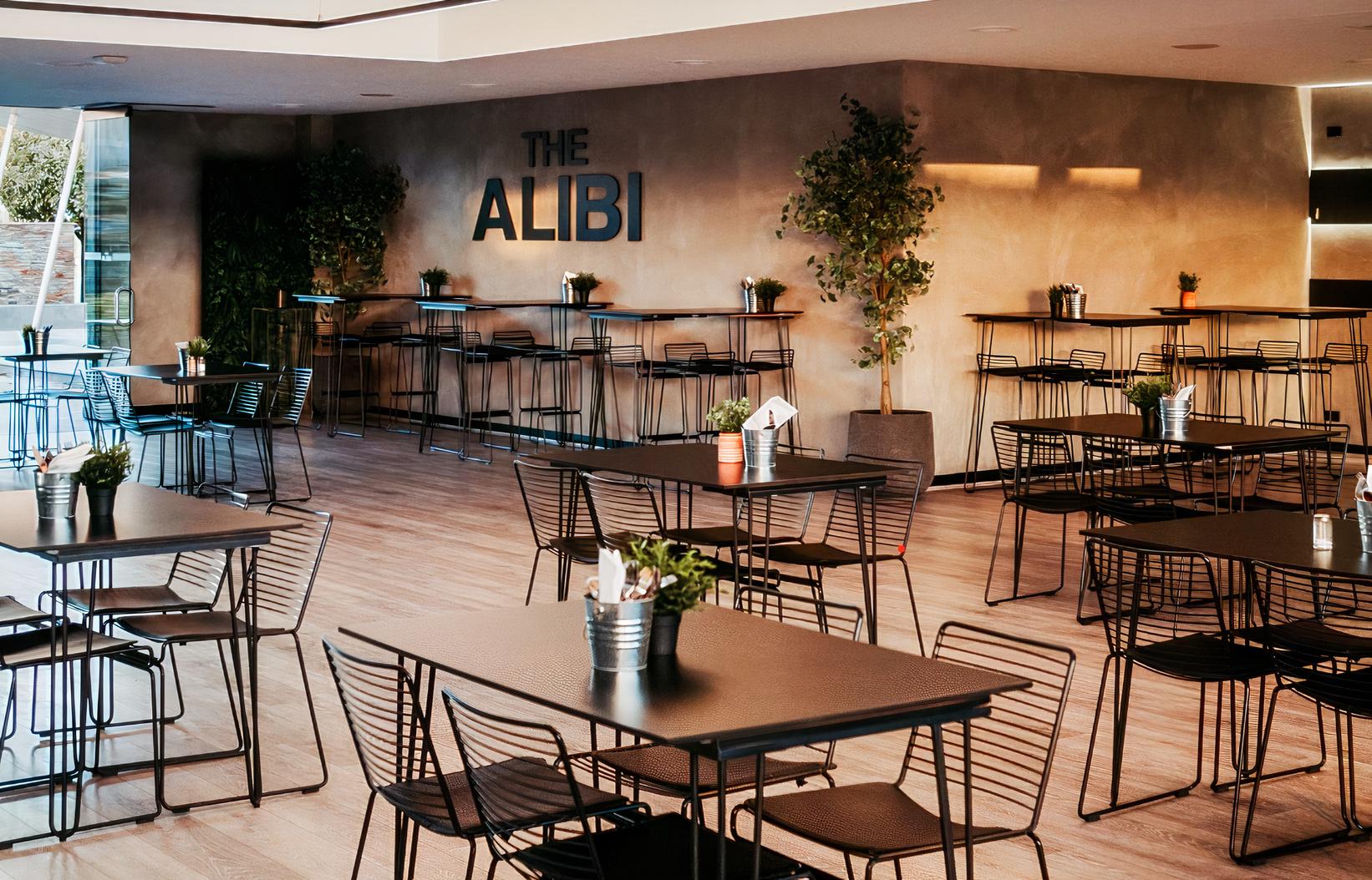 A cafe with metal tables and chairs, potted plants, and a light wood floor at The Alibi.
