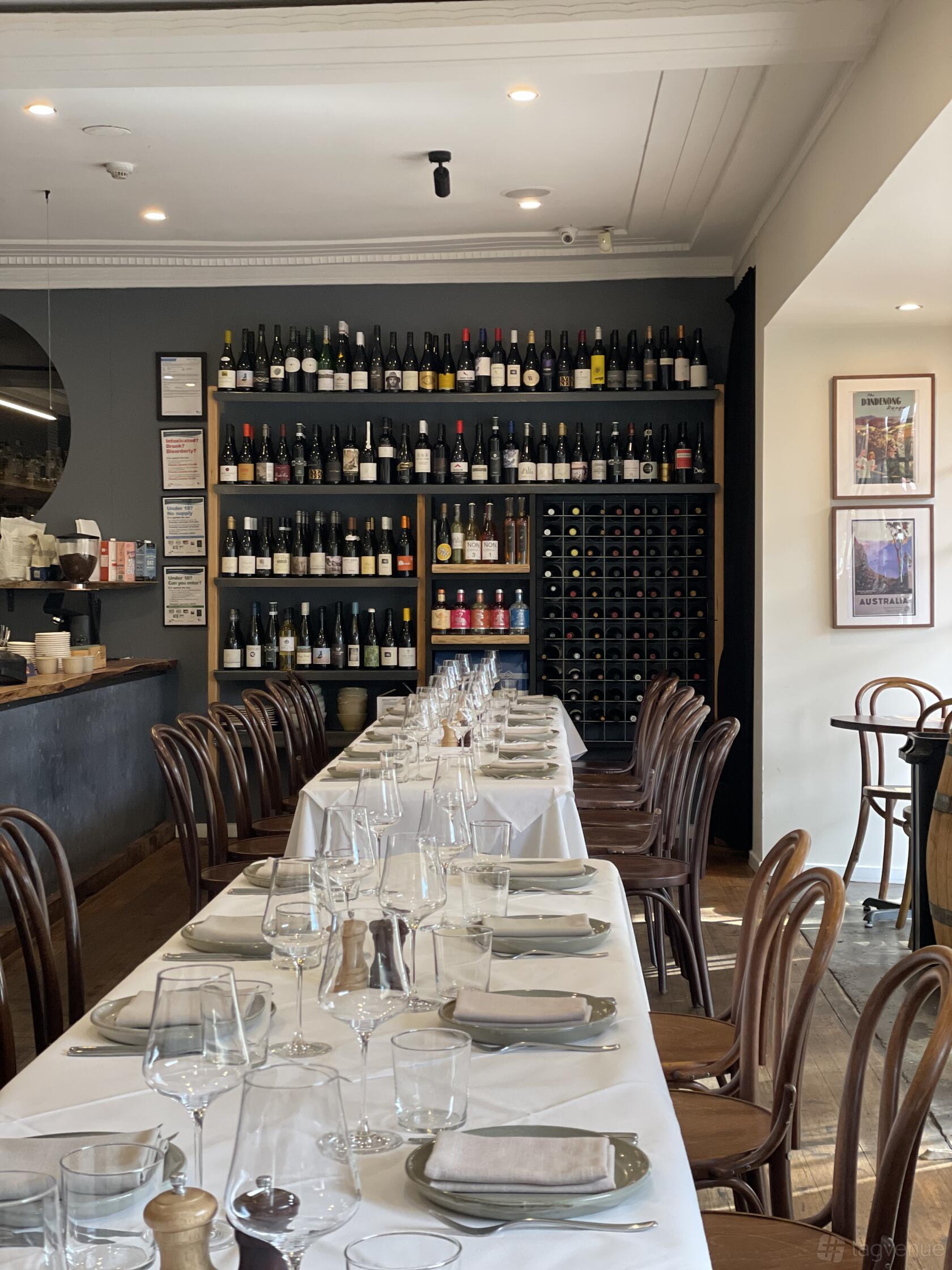 A restaurant dining area with a long table set with glassware and white linens, surrounded by shelves of wine bottles at Cognoscenti Food & Wine.
