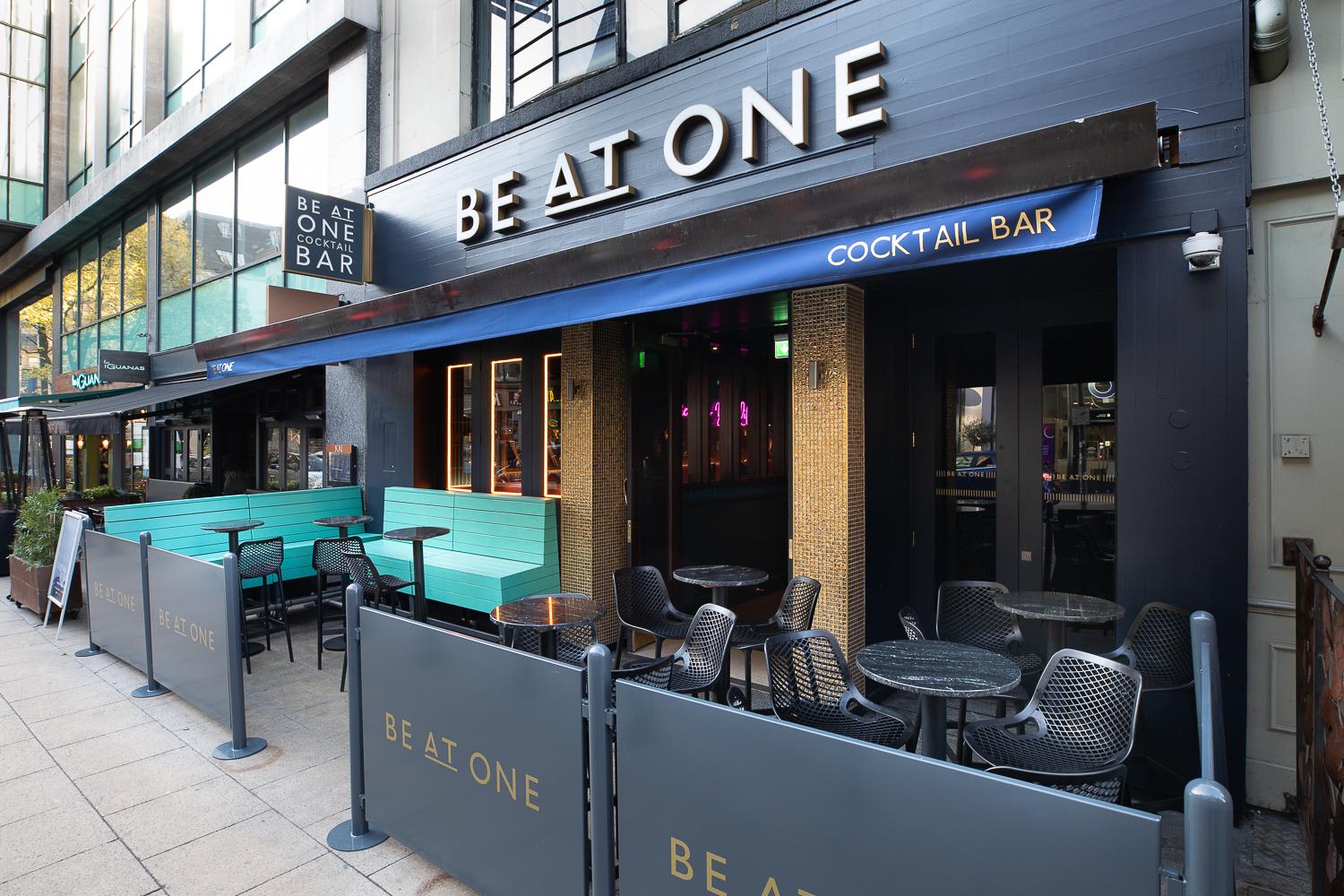 A pub with outdoor seating, turquoise benches, and round tables under a blue awning at Be At One Manchester.