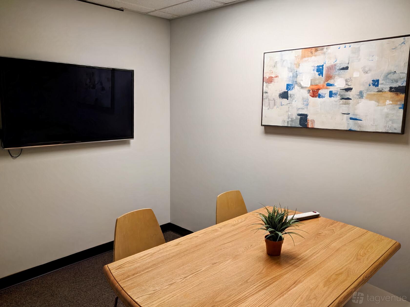 A meeting room with a wooden table, two chairs, a wall-mounted TV, and abstract artwork at West Seattle Coworking.