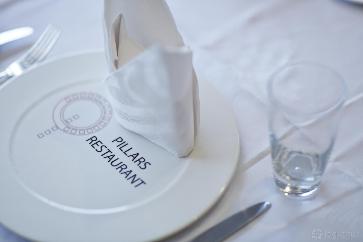 A table setting with a neatly folded napkin, branded charger plate, and glass at Pillars Restaurant, University of West London.