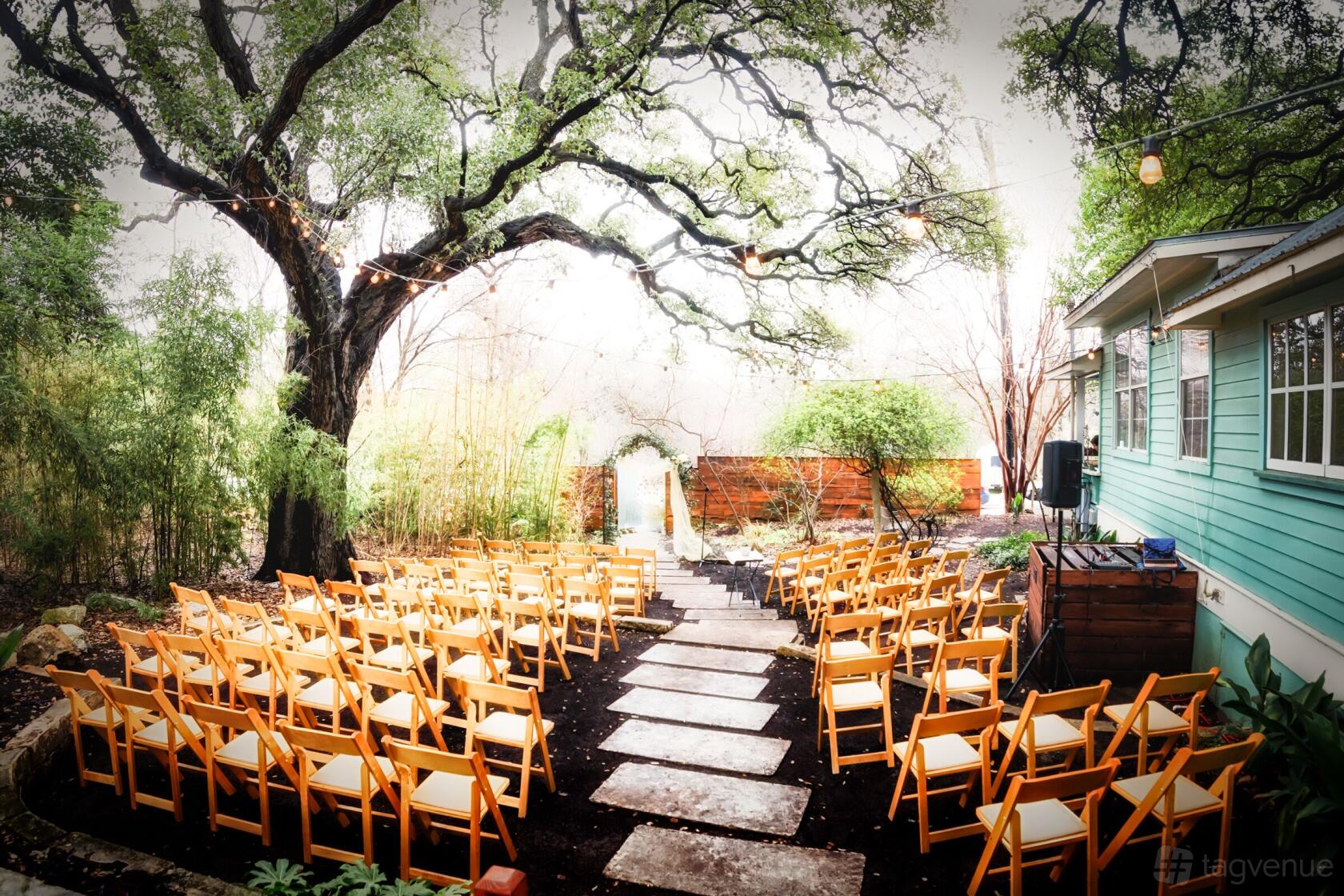An outdoor event venue with rows of wooden chairs, stone walkway, and large oak trees at Sanctuary Event Space.