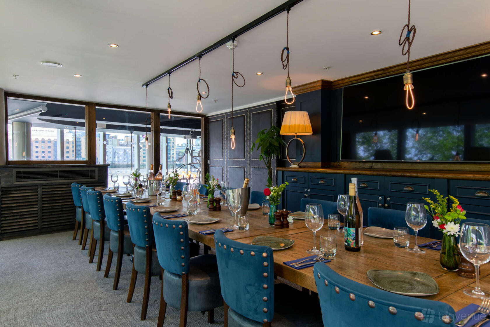 A pub dining room with a long wooden table, blue velvet chairs, and hanging Edison bulbs at The Oyster Shed.