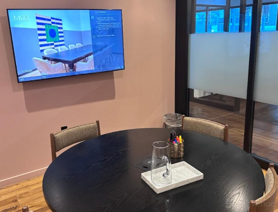 A meeting room with a round black table, chairs, wall-mounted screen, and glass door at Mindspace Wynwood.